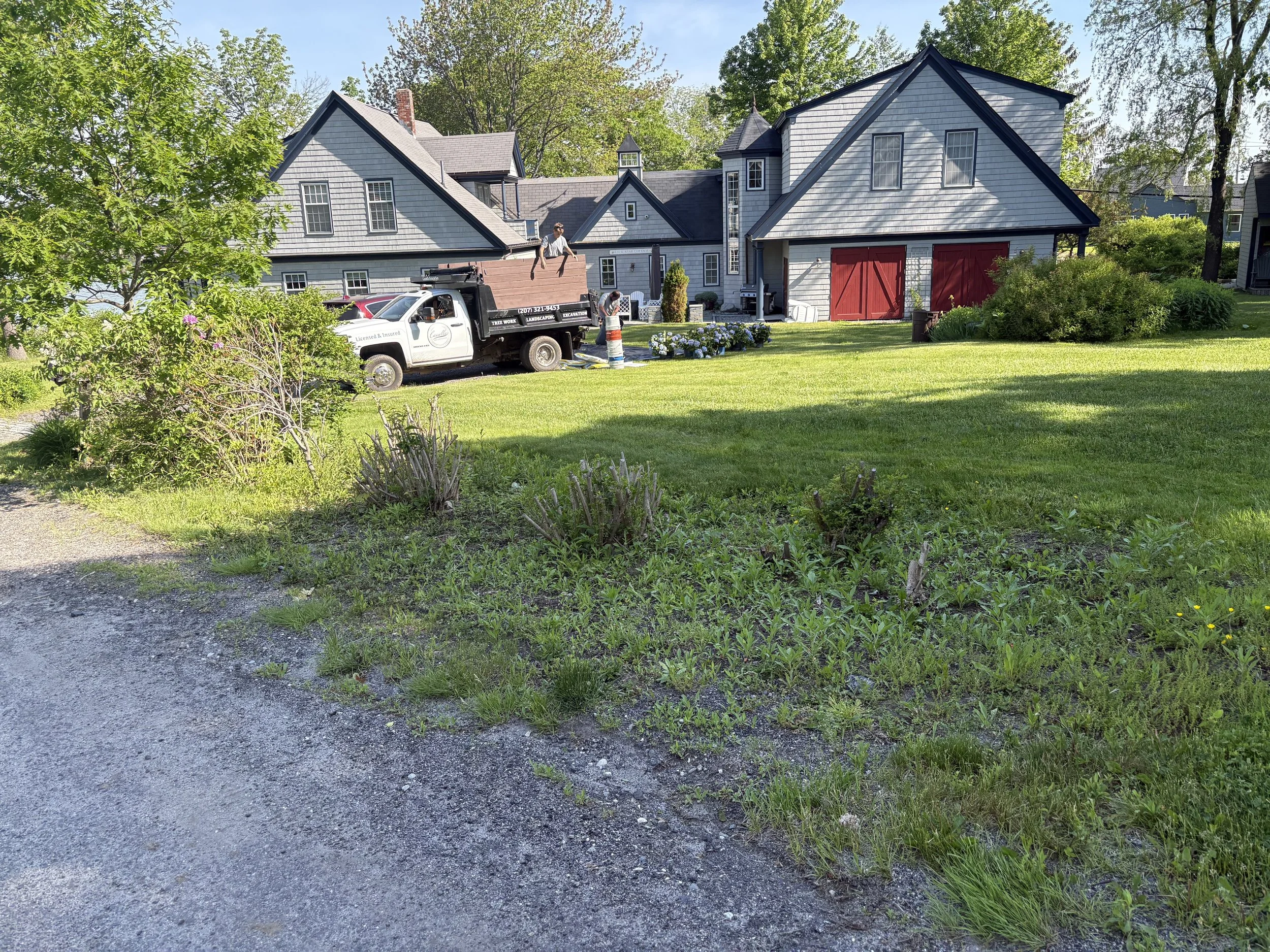 A house with gray siding and a gray roof, red doors on the garage, surrounded by greenery and trees, with a lawn and flowerbeds. Construction or landscaping workers near a white truck are working.
