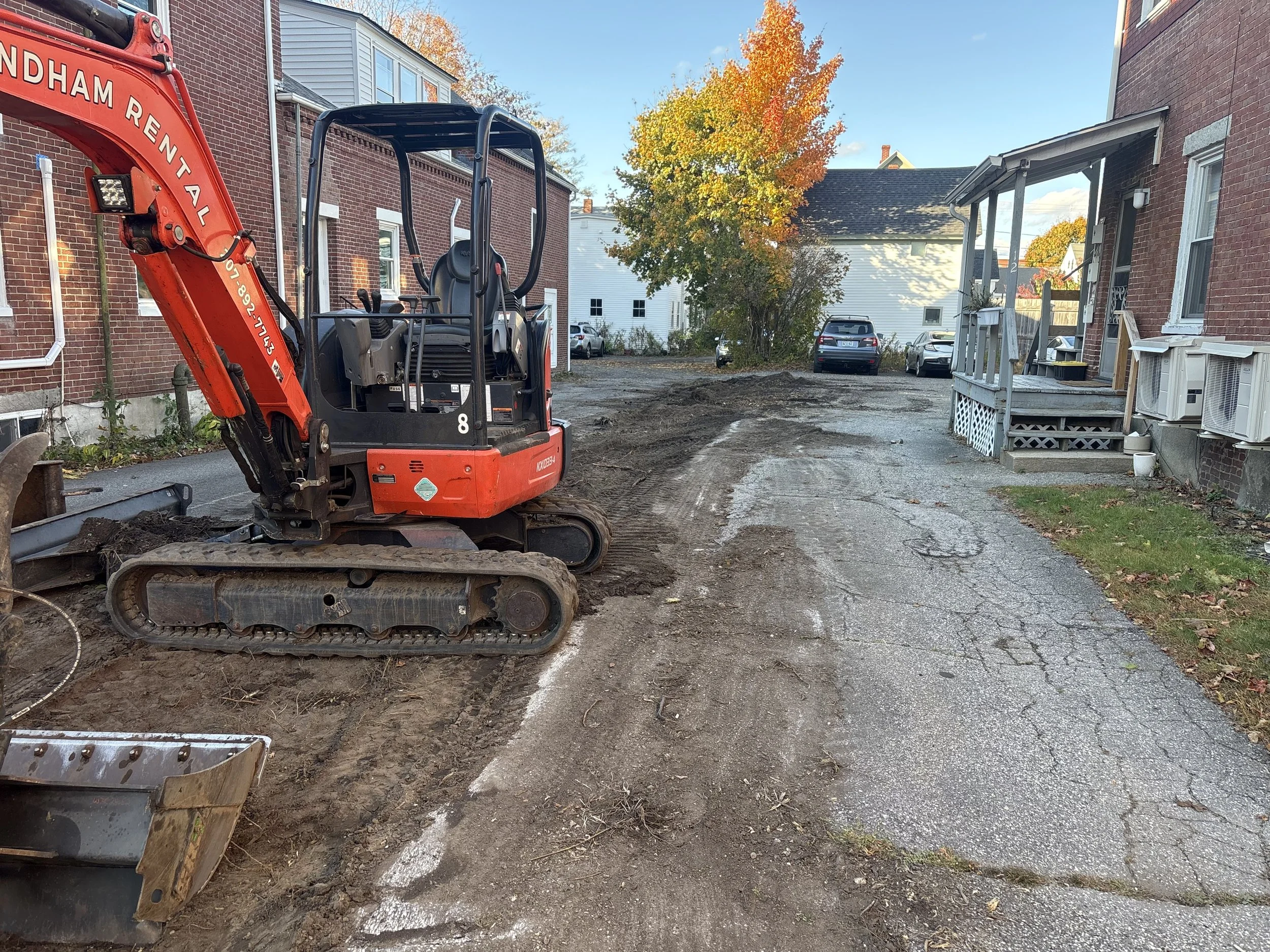 A small excavator parked on a dirt driveway next to a brick building with parked cars in the background and autumn trees with orange and yellow leaves.