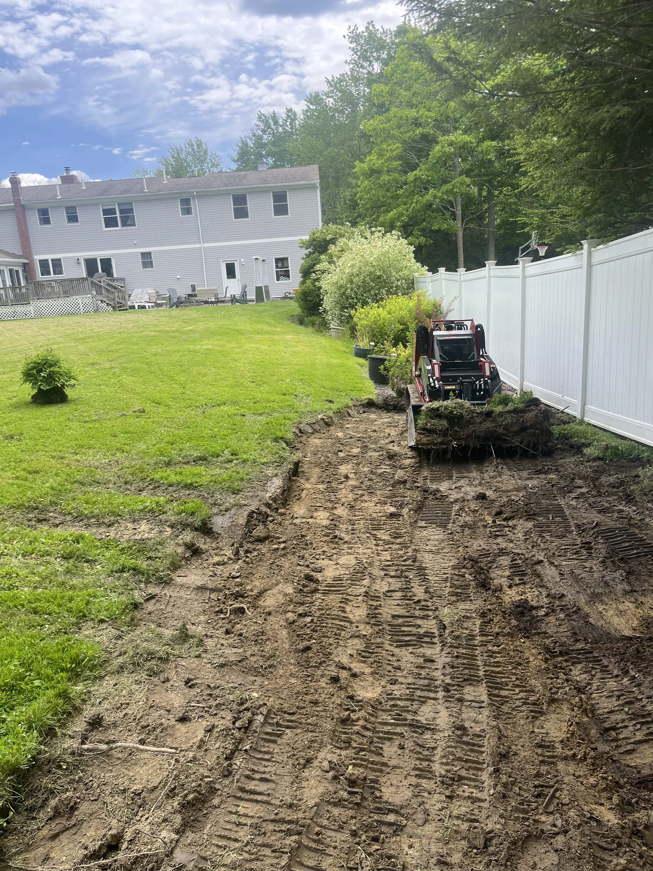 A backyard with a green lawn, bushes, and trees, fenced with a white privacy fence, and a garden tractor on a dirt path where a trench is being dug.
