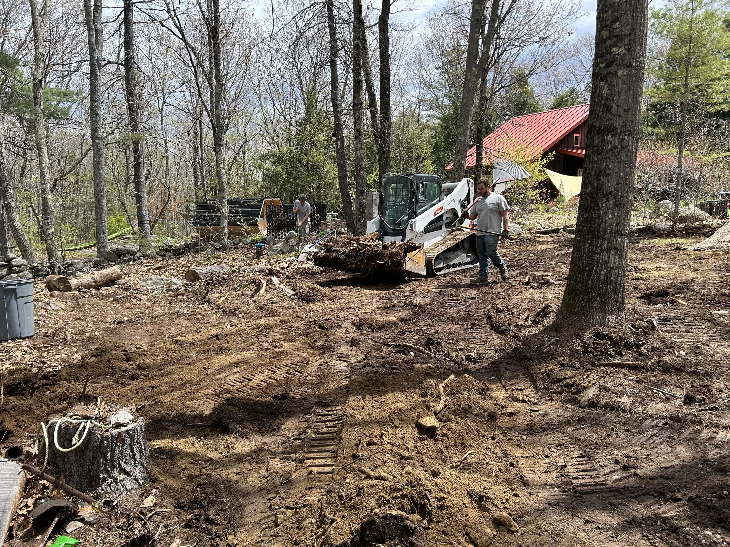 Two workers operate a small excavator on a cleared, rocky, wooded area near a red barn, with tree stumps and construction equipment visible.