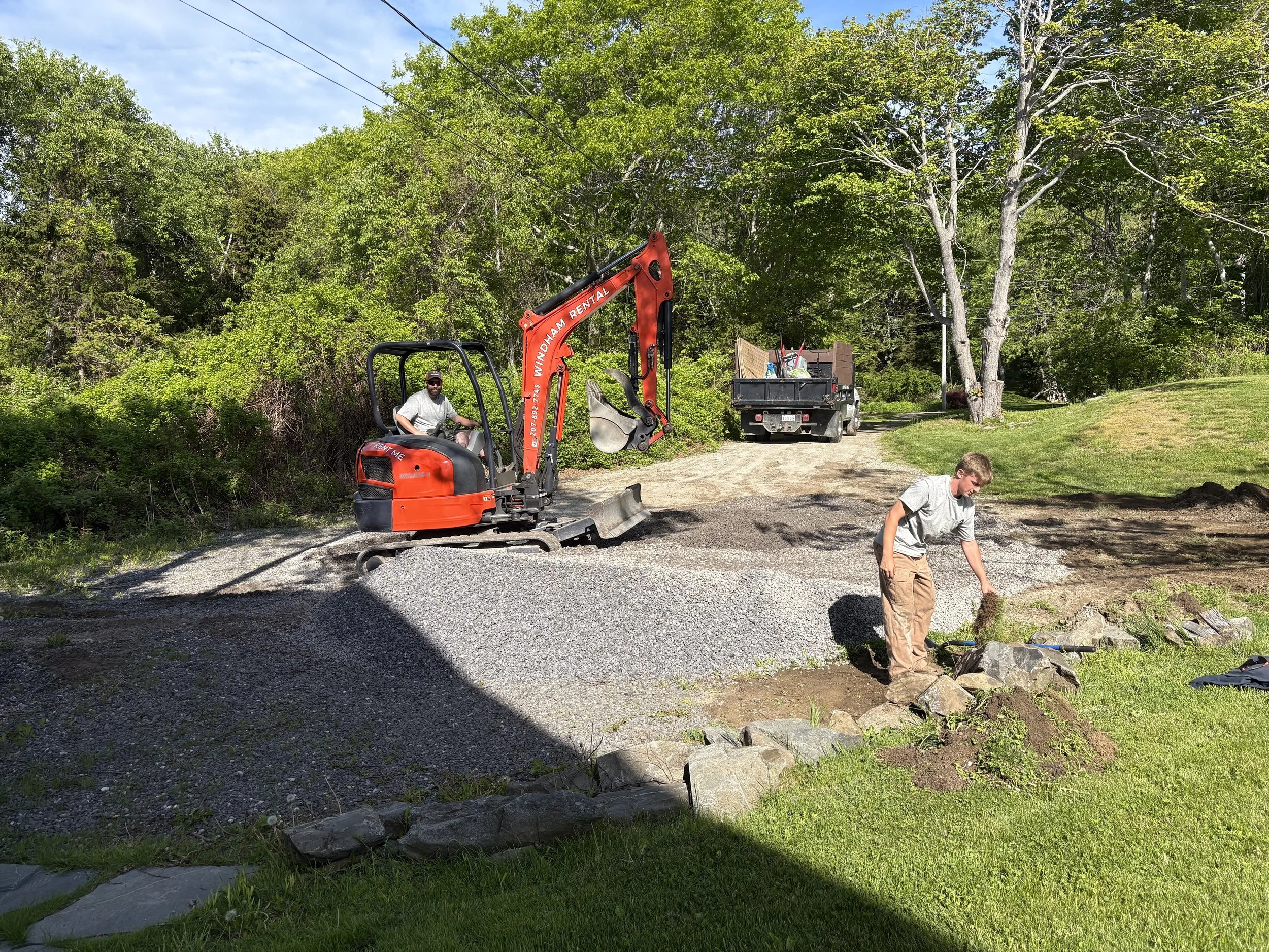 People working on a landscaping project with a mini excavator on a dirt path surrounded by trees and grass.