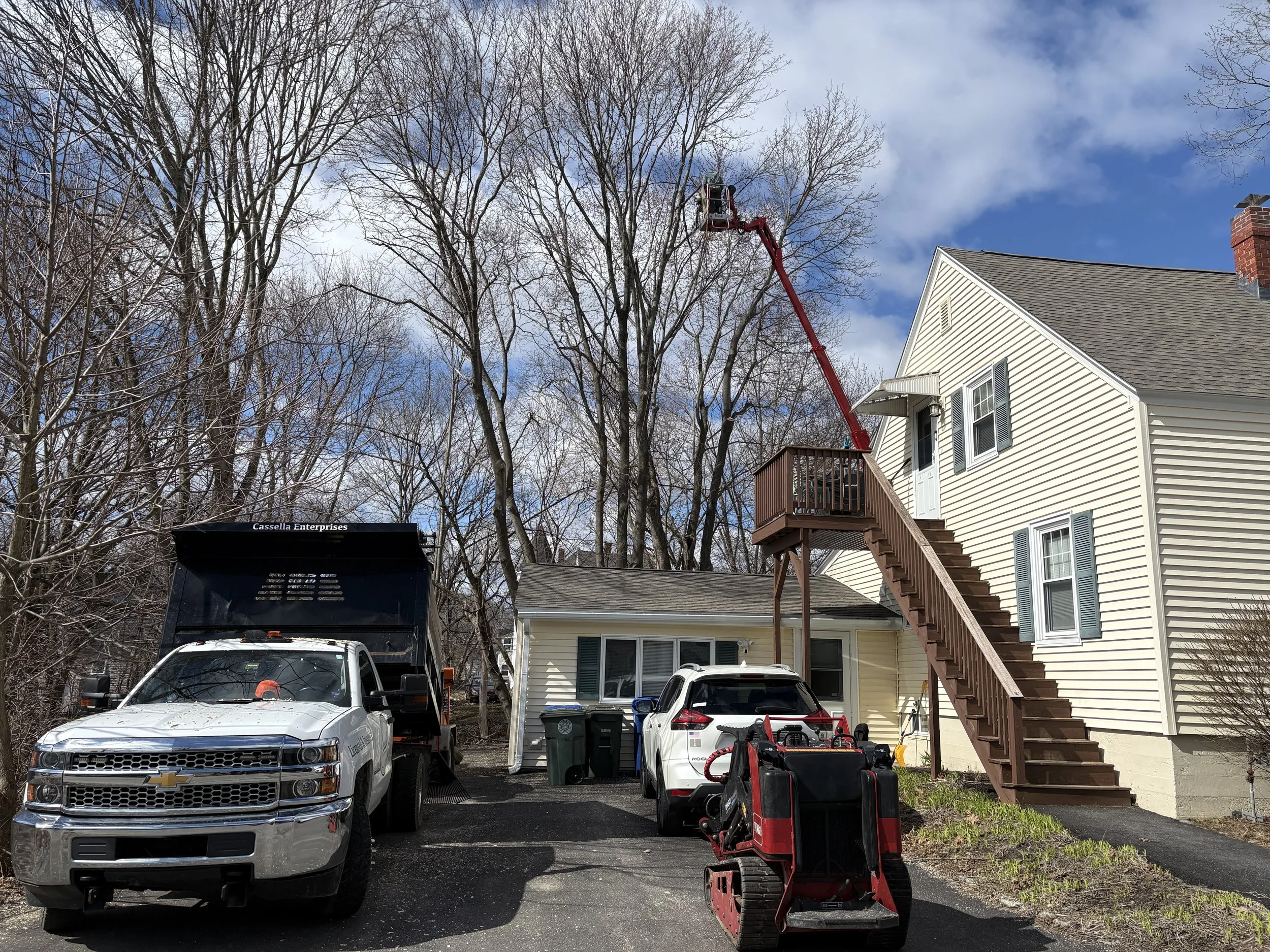 A residential backyard with a two-story white house featuring a wooden staircase, and vehicles parked in the driveway. Tree branches with no leaves are visible, and a cherry picker lift is extending up into the trees.