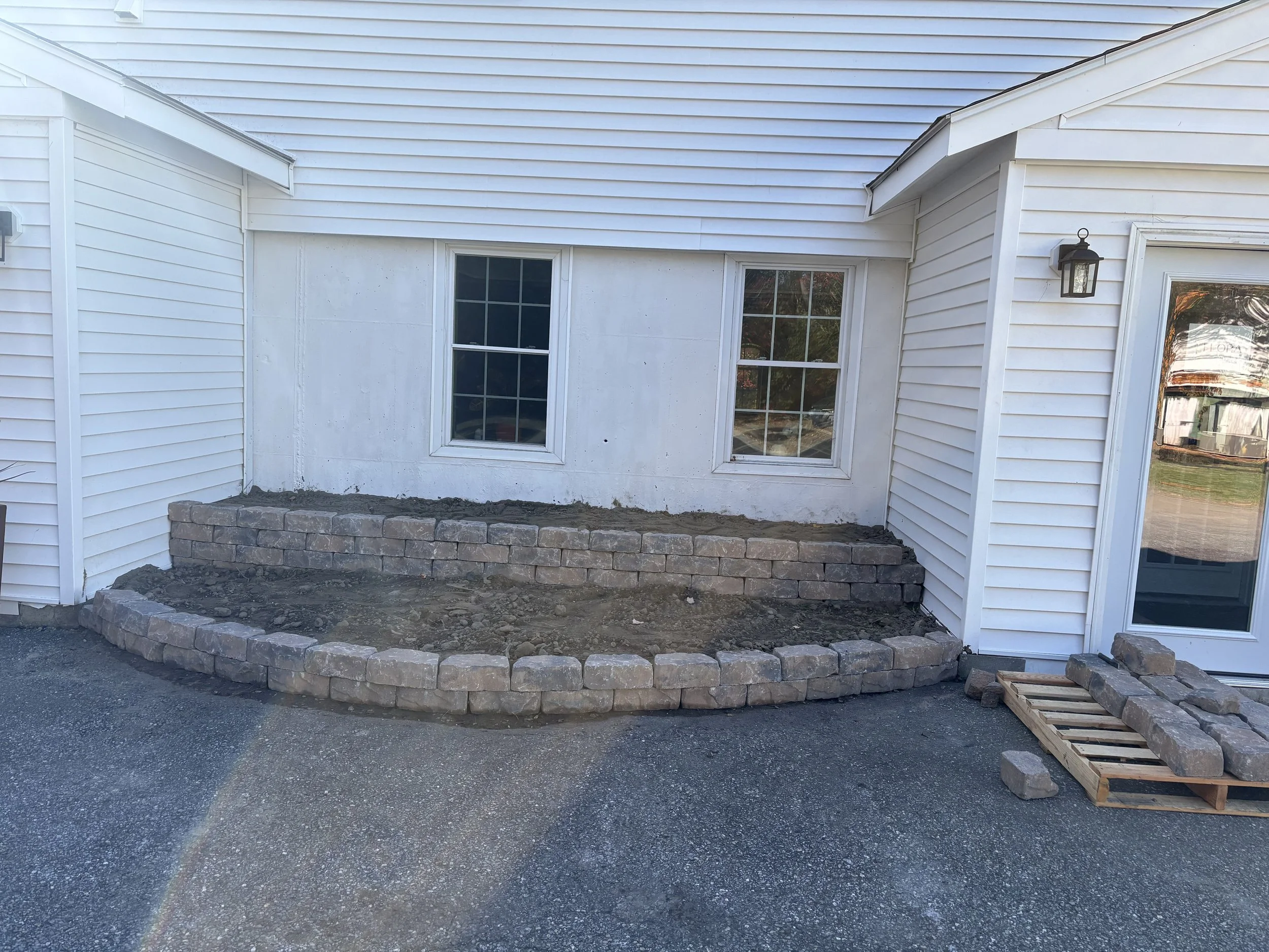 White house exterior with two windows, a porch light, and an ongoing landscaping project with a brick border wall under construction.