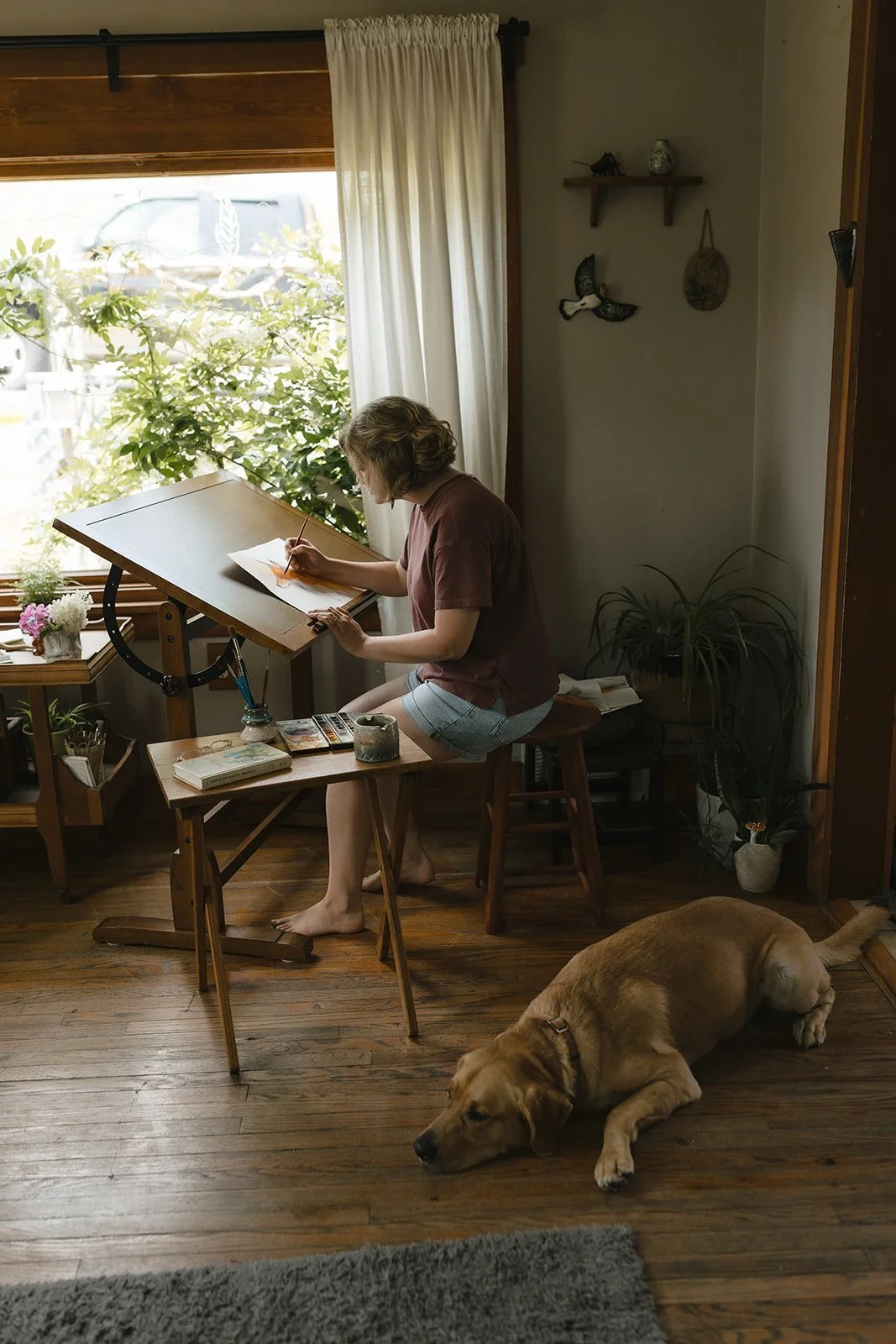 artist painting at an easel while a dog sleeps by her feet.
