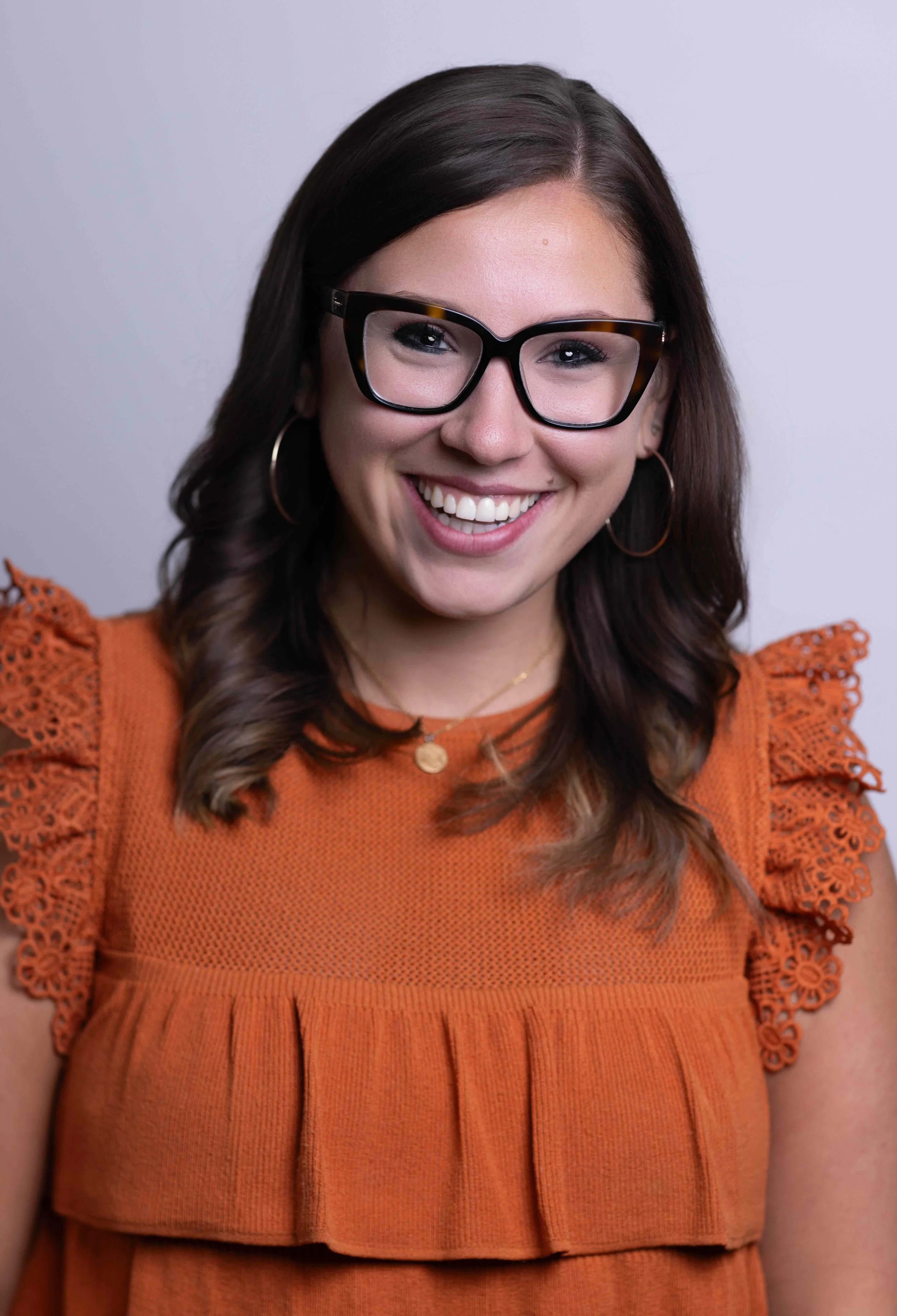 Close-up of a smiling woman with long brown hair, wearing large black glasses, gold hoop earrings, a gold necklace, and an orange dress with lace details on the shoulders.