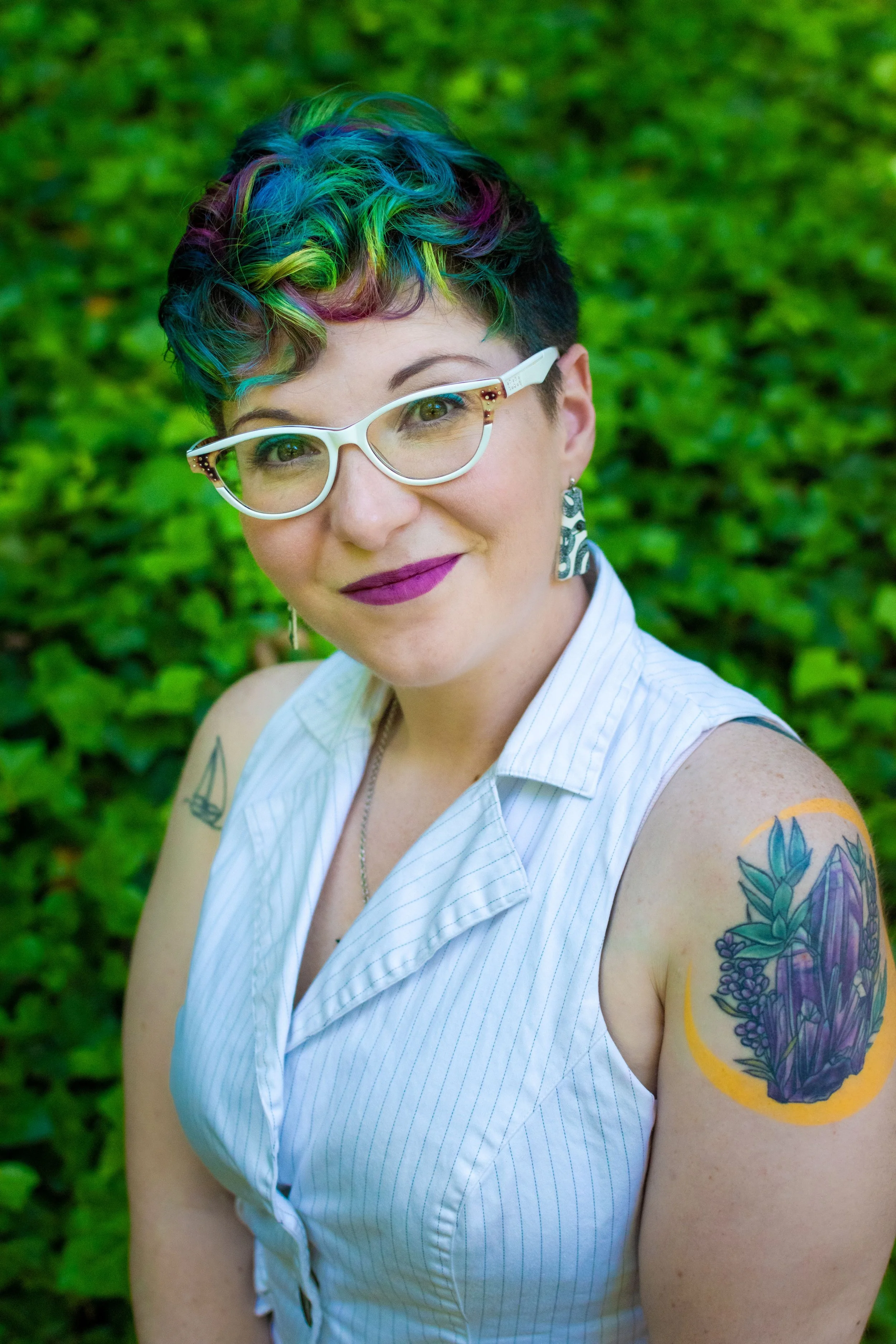 A woman with short, curly hair dyed in rainbow colors, wearing white glasses, earrings, wearing a sleeveless, white, pinstriped blouse, and displaying a colorful floral tattoo on her upper arm. She is standing outdoors with a blurred green foliage background.