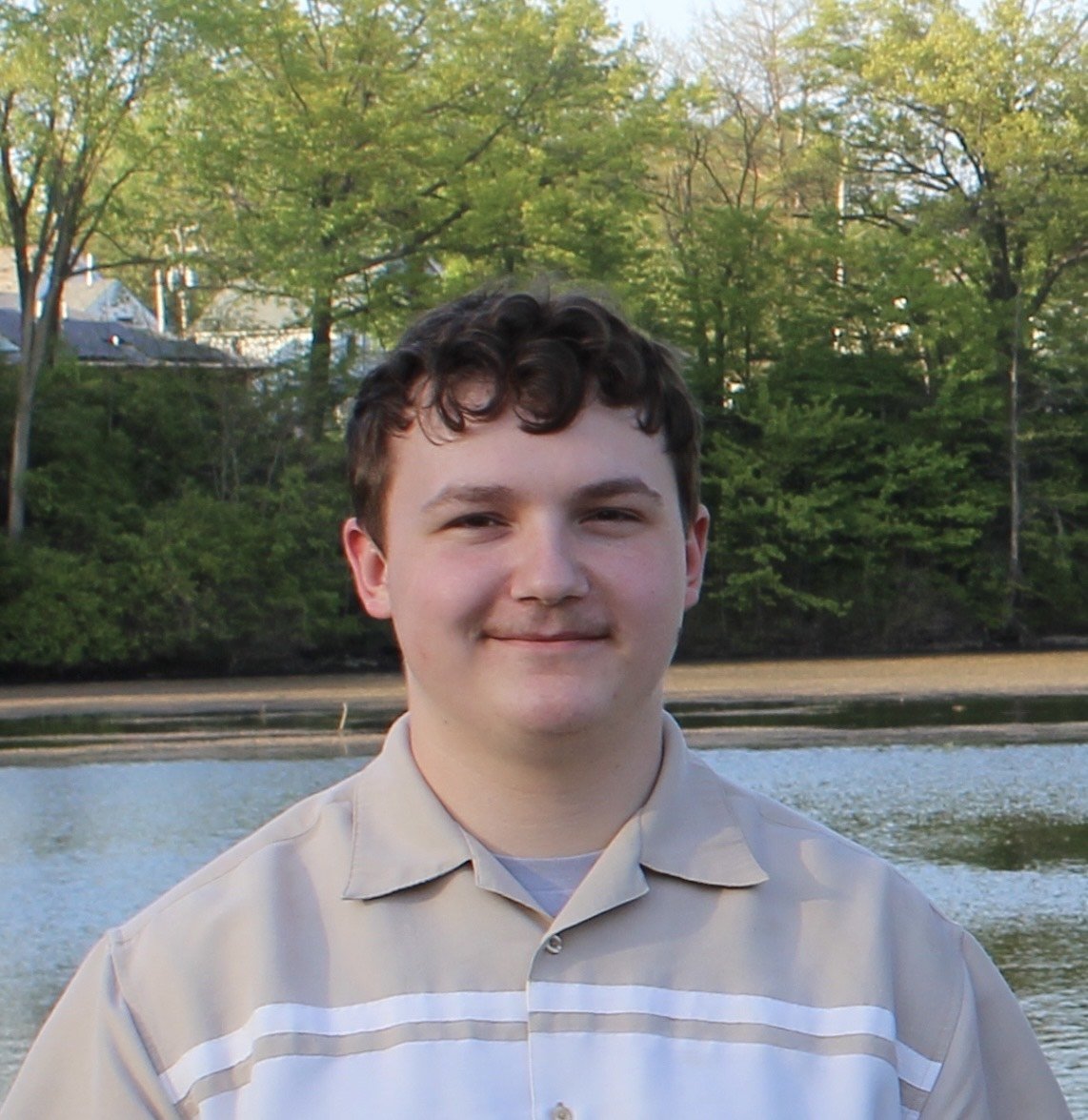 A young man with short curly brown hair and light skin smiling outdoors near a body of water, with green trees and a clear sky in the background.