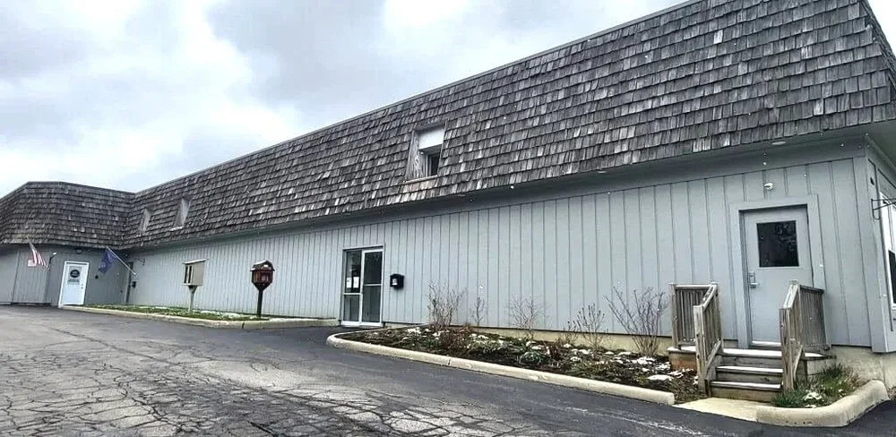 A long, light gray building with a steep, shingled roof and a few small windows. There are steps leading up to a side door, a small parking lot in front, and a mailbox near the entrance. Flags and a flagpole are visible on the left side of the building.