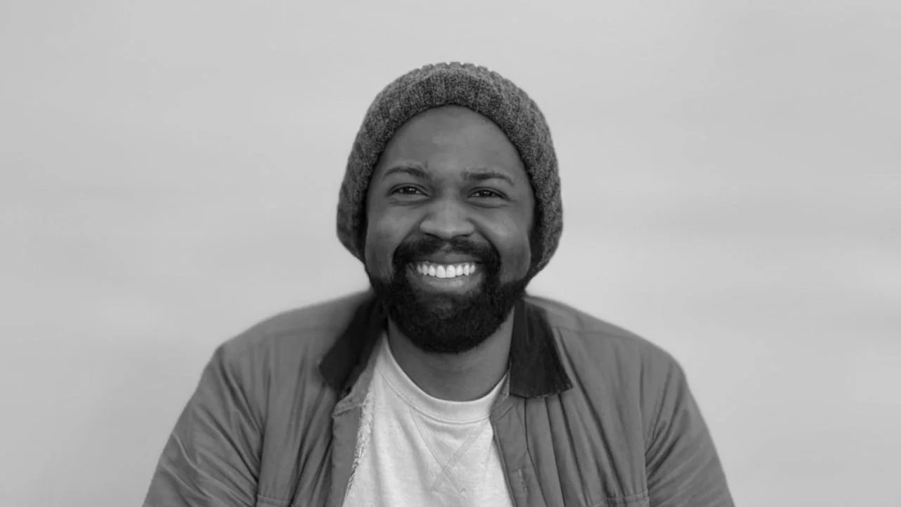 Black man smiling, wearing a gray beanie, a jacket, and a light-colored shirt, against a plain background.