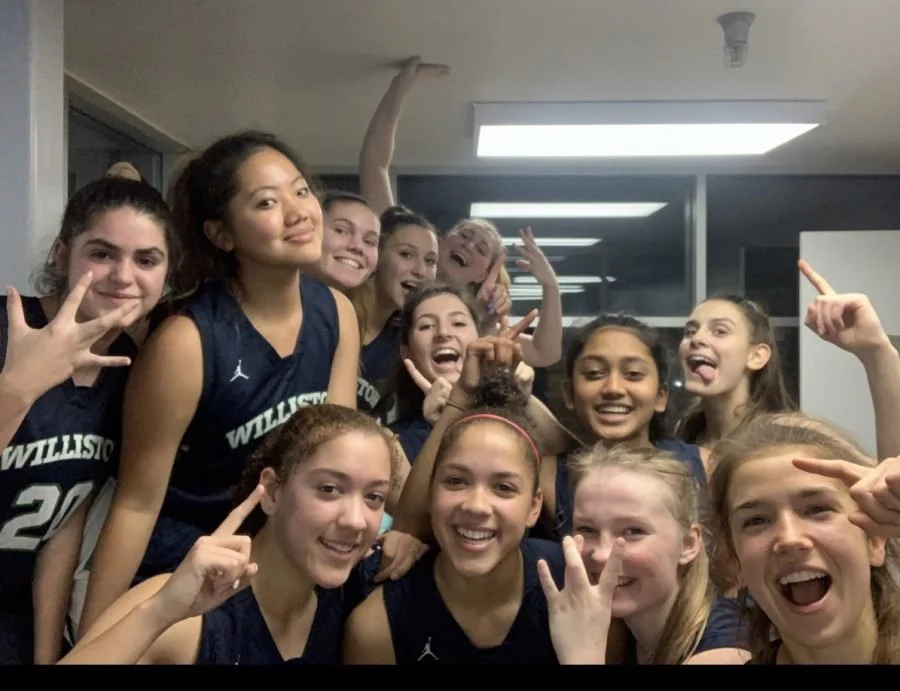 Group of young female basketball players in Williston jerseys celebrating and posing together indoors.