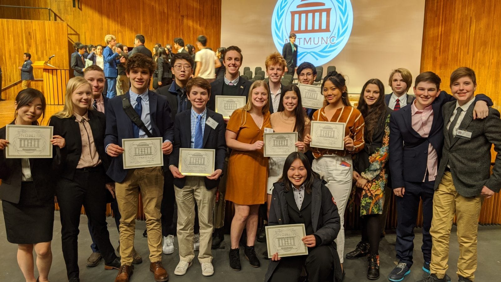 Group of young students with diplomas at an educational award ceremony, standing on stage in front of a large screen showing a logo with a building and laurel wreath.