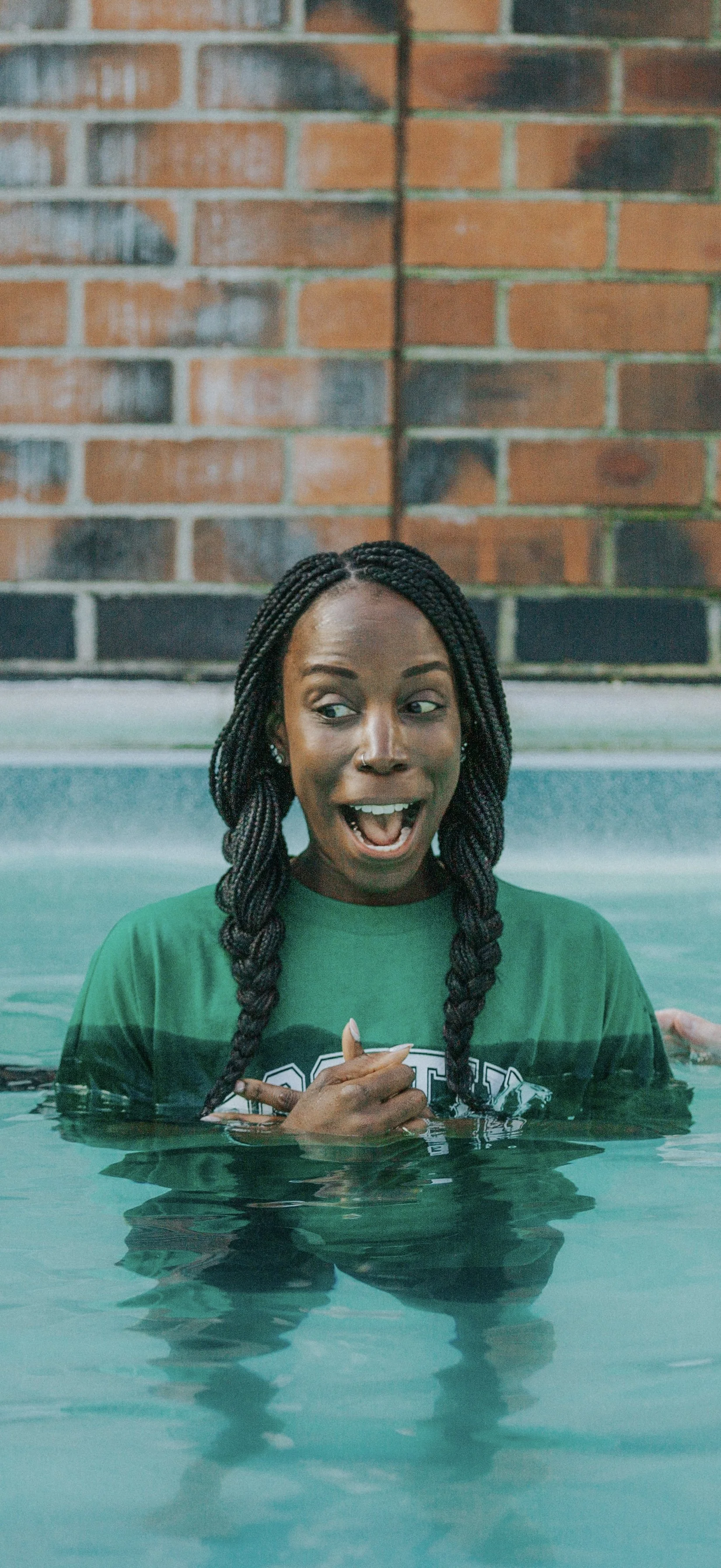 A woman with braided hair and a green shirt in a swimming pool, smiling and pointing at herself, with a brick wall background.