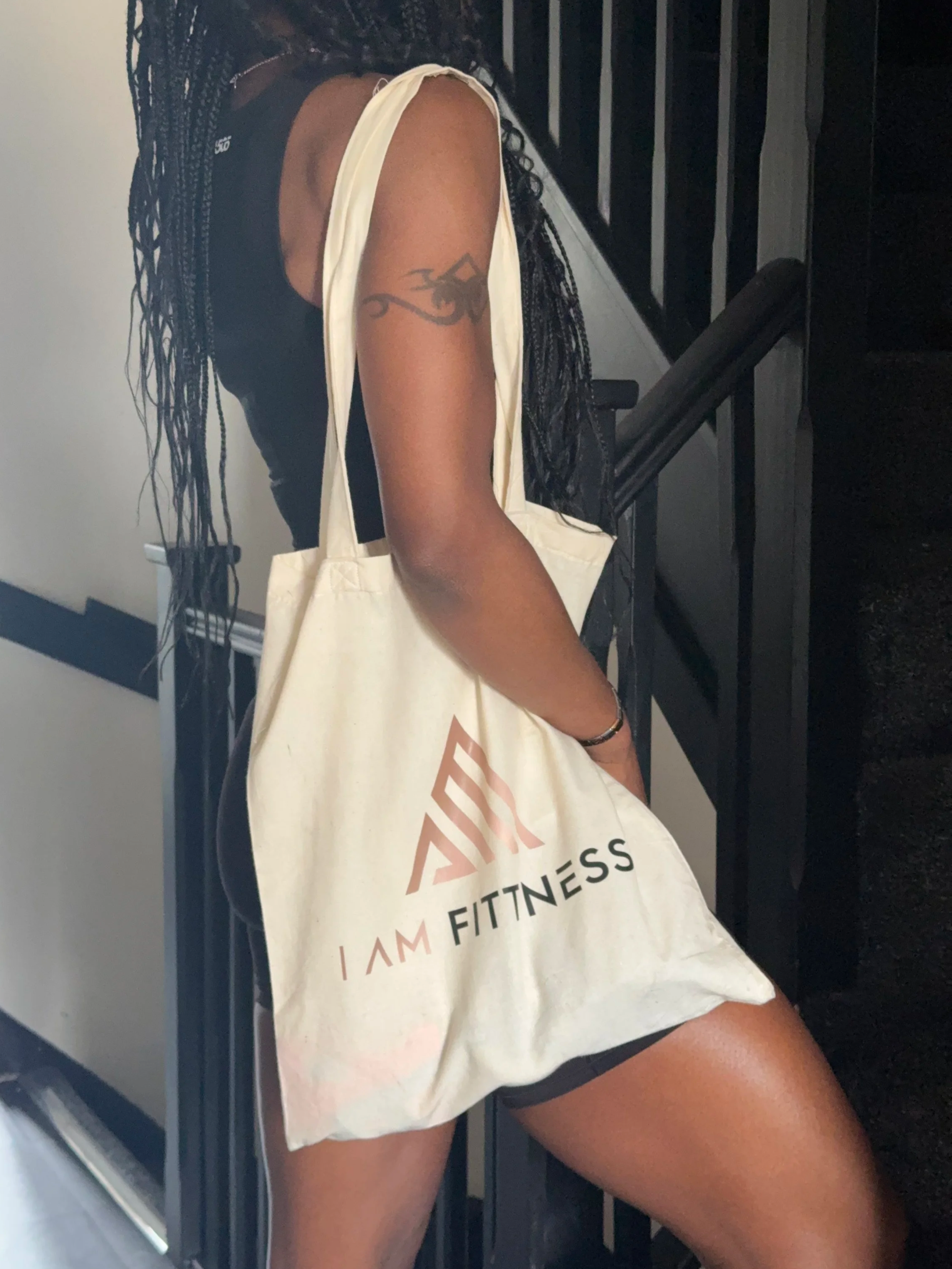 A woman with long braided hair standing near a staircase, wearing a black sleeveless top and black shorts, carrying a white tote bag with "I AM FITNESS" printed on it.