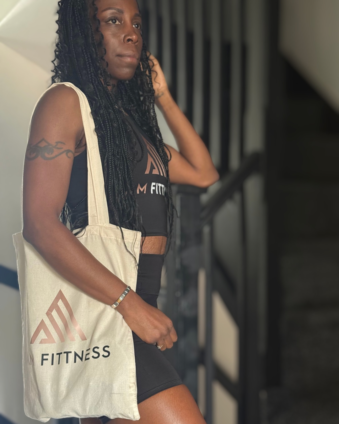 A young woman with black braided hair standing indoors near a staircase, wearing a black athletic top and shorts, carrying a beige tote bag with a logo and the words 'FITNESS' on it, and wearing a bracelet.