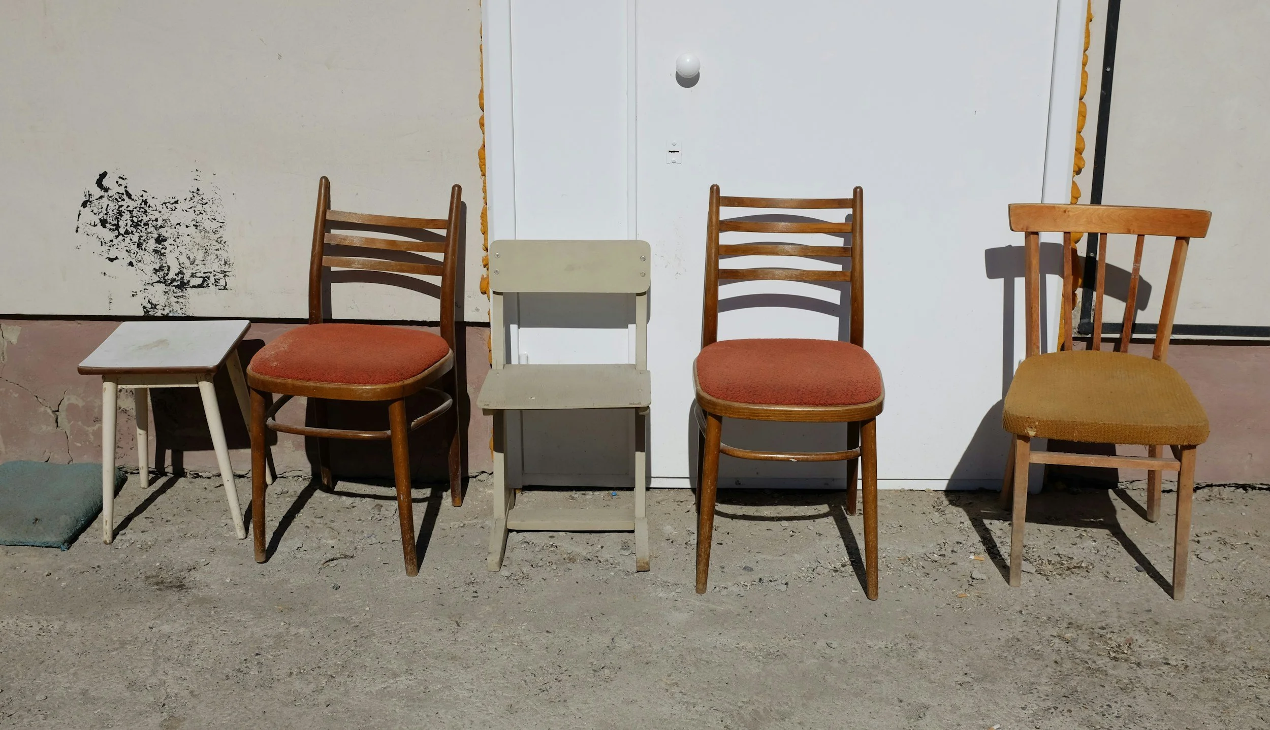 Five chairs of different styles and colors lined up outside, with a white door behind them. The ground is dirt, and there is some black paint splash on the wall to the left.