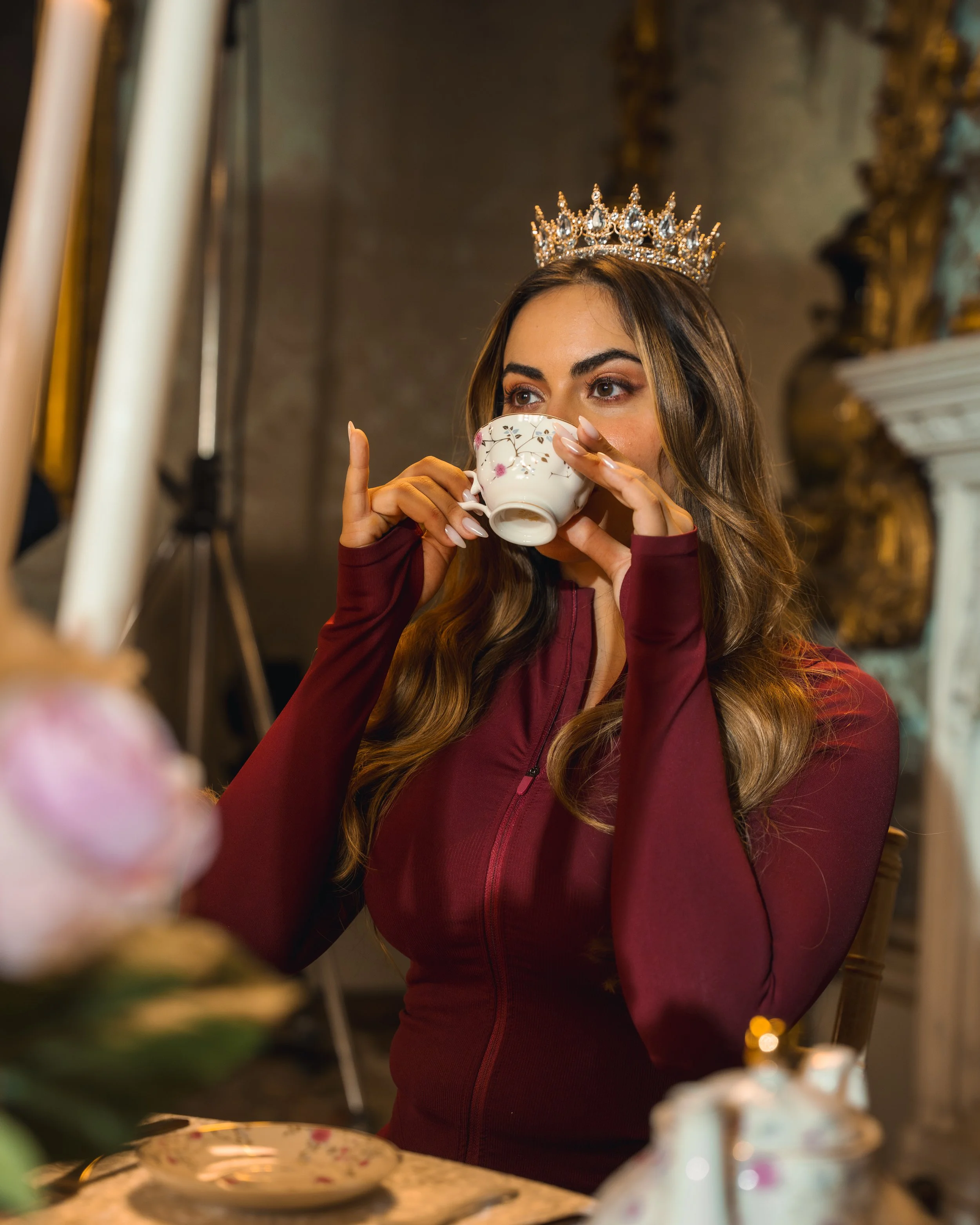 A woman wearing a crown drinking from a teacup in a decorated room.