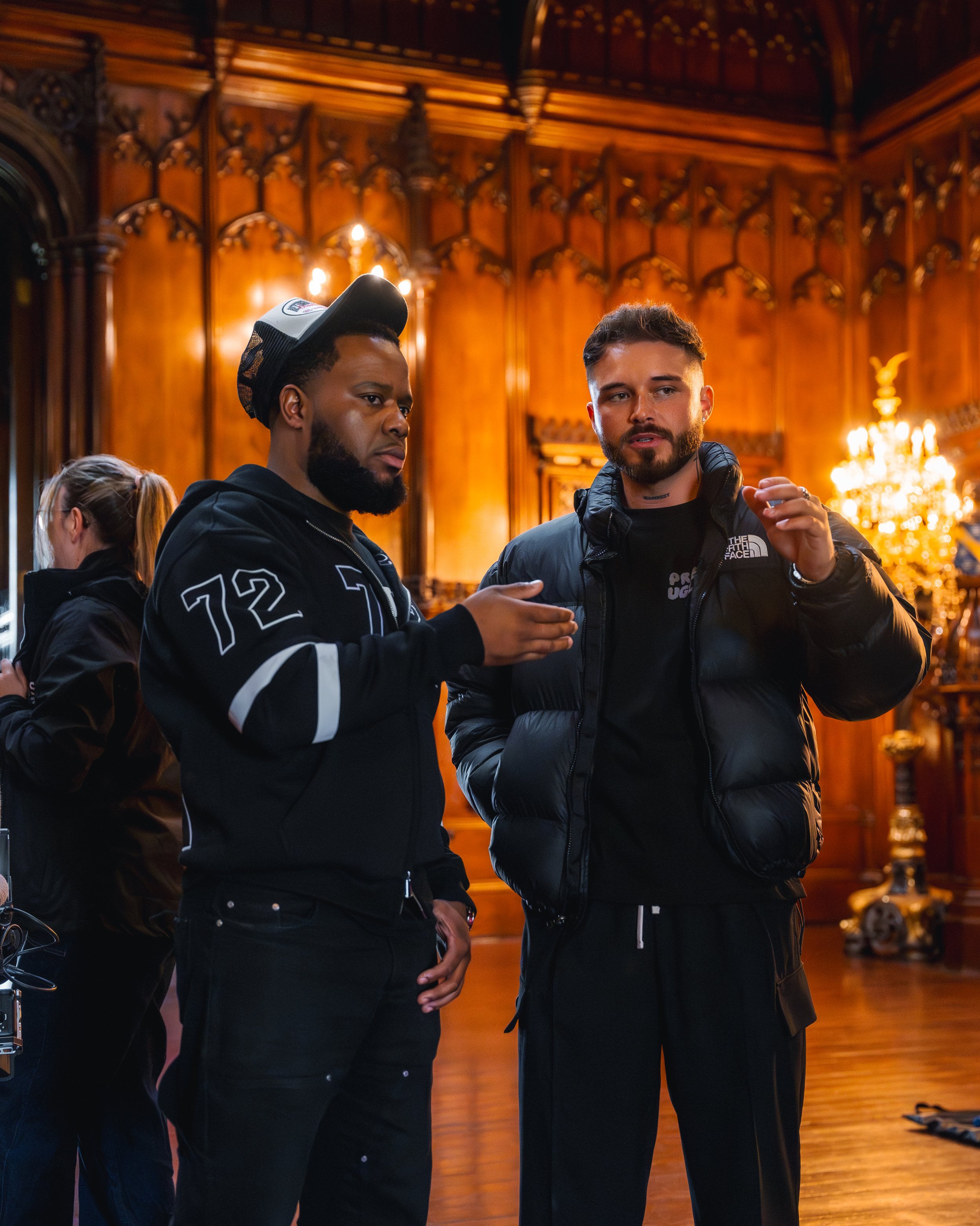Two men engaged in conversation in a richly decorated wood-paneled room with warm lighting and a chandelier in the background.