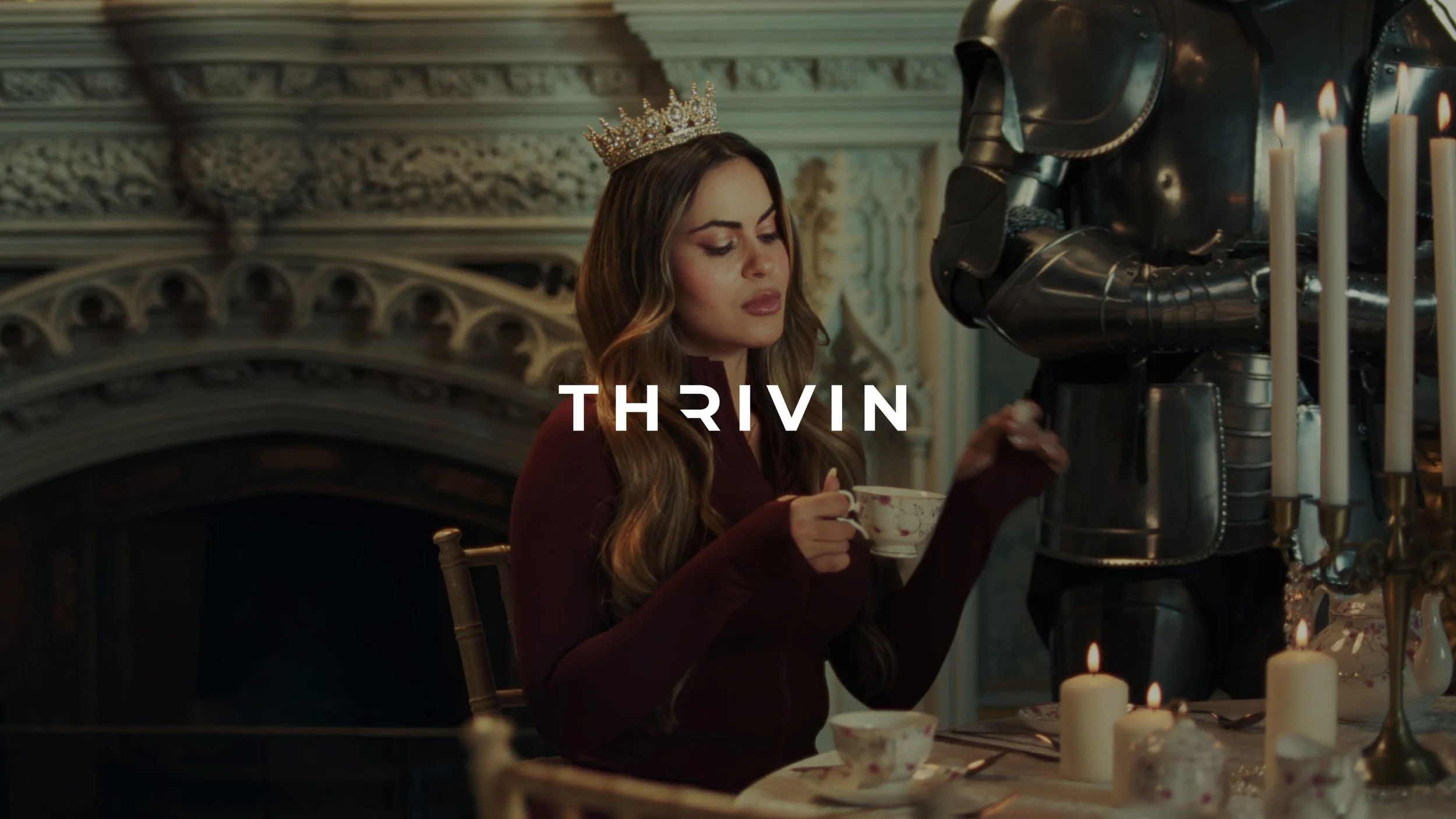 Woman with long wavy hair wearing a crown, holding a teacup, sitting at a decorated table in a lavish room, with a figure in a suit of armor on her right, and lit candles on the table.