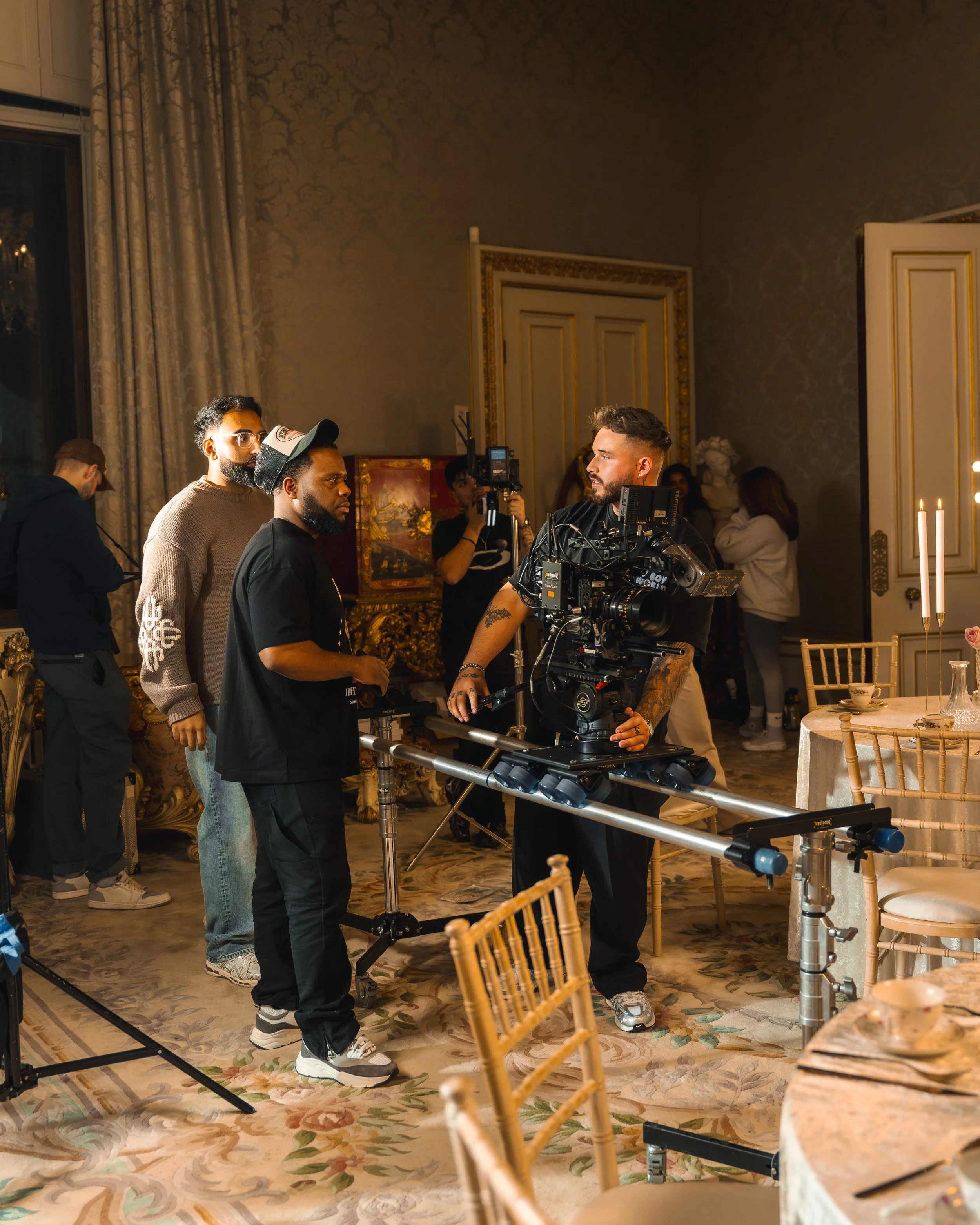 Filmmaking crew setting up camera on tripod in elegant room with round table and gold chairs.