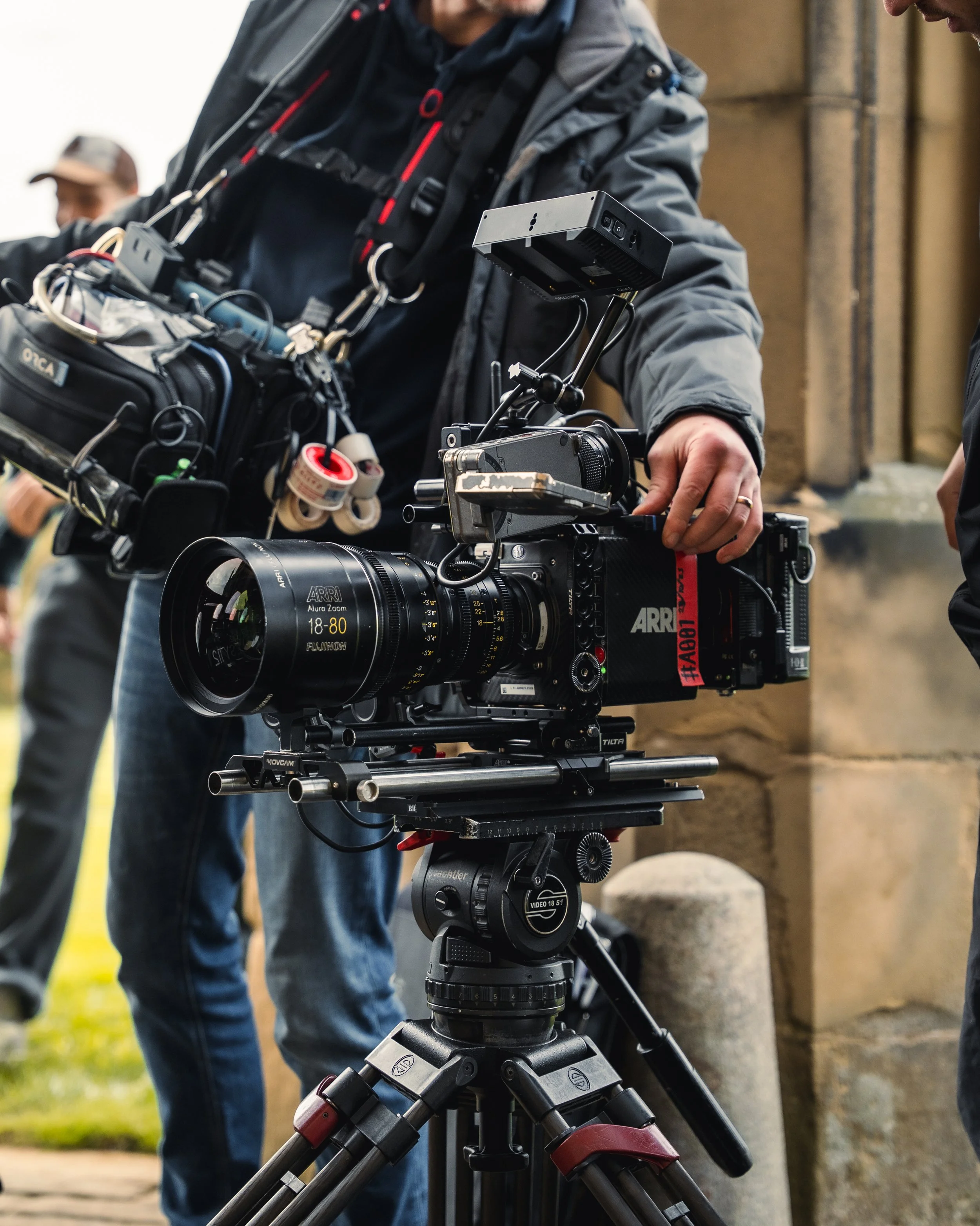 Close-up of a professional movie camera on a tripod being operated by a crew member with a gray jacket, with another crew member visible in the background.