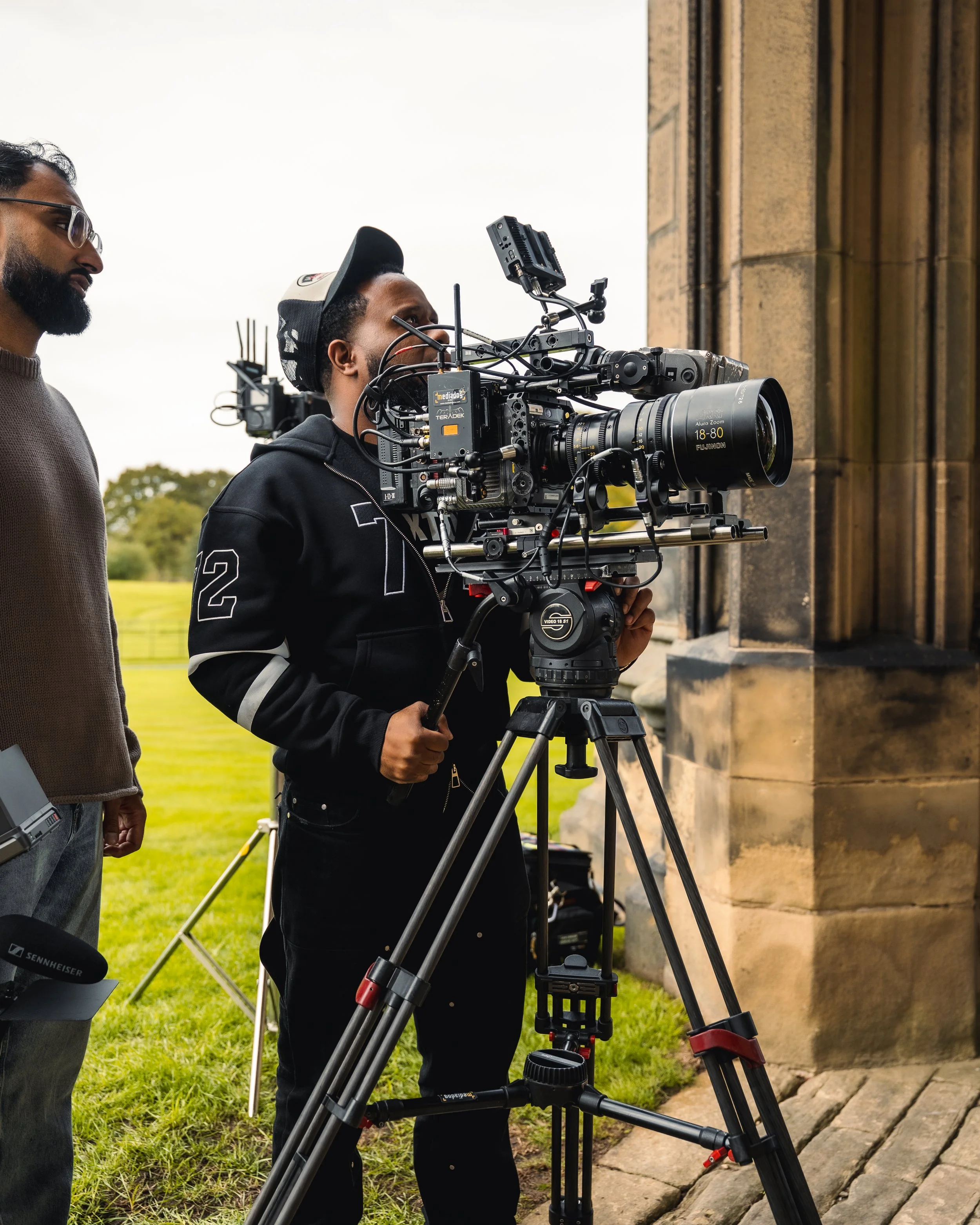 Two men filming with a professional large camera mounted on a tripod outside a stone building, with green grass and trees in the background.