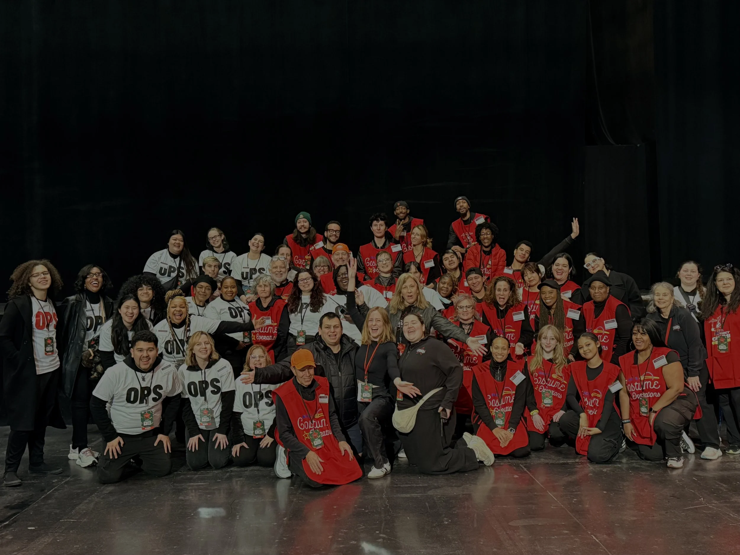 A large group of diverse people posing together on stage, some in red vests, some in white shirts, smiling and making various gestures.