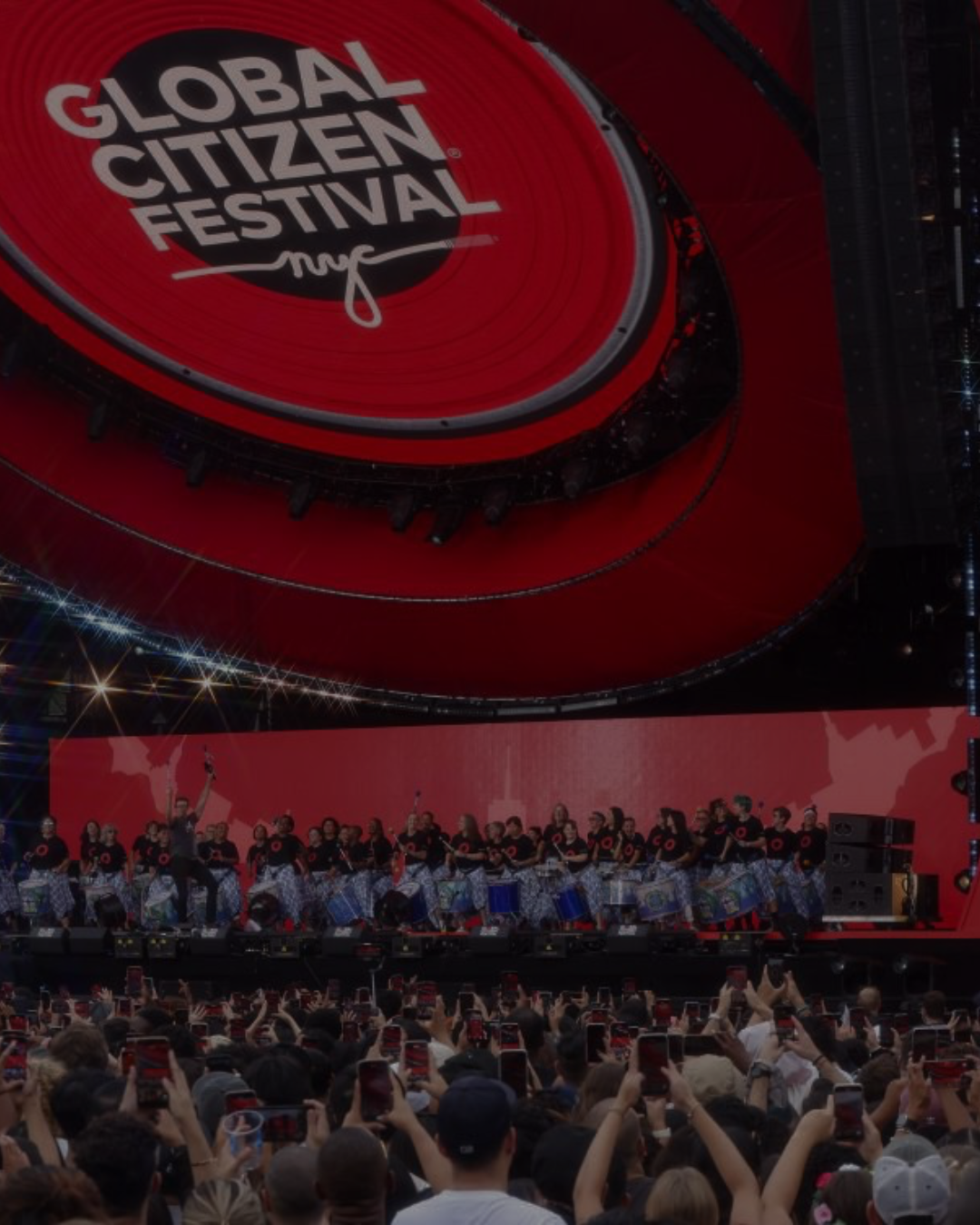 A large crowd watching a live performance at the Global Citizen Festival with a stage full of performers and a big circular digital screen overhead.