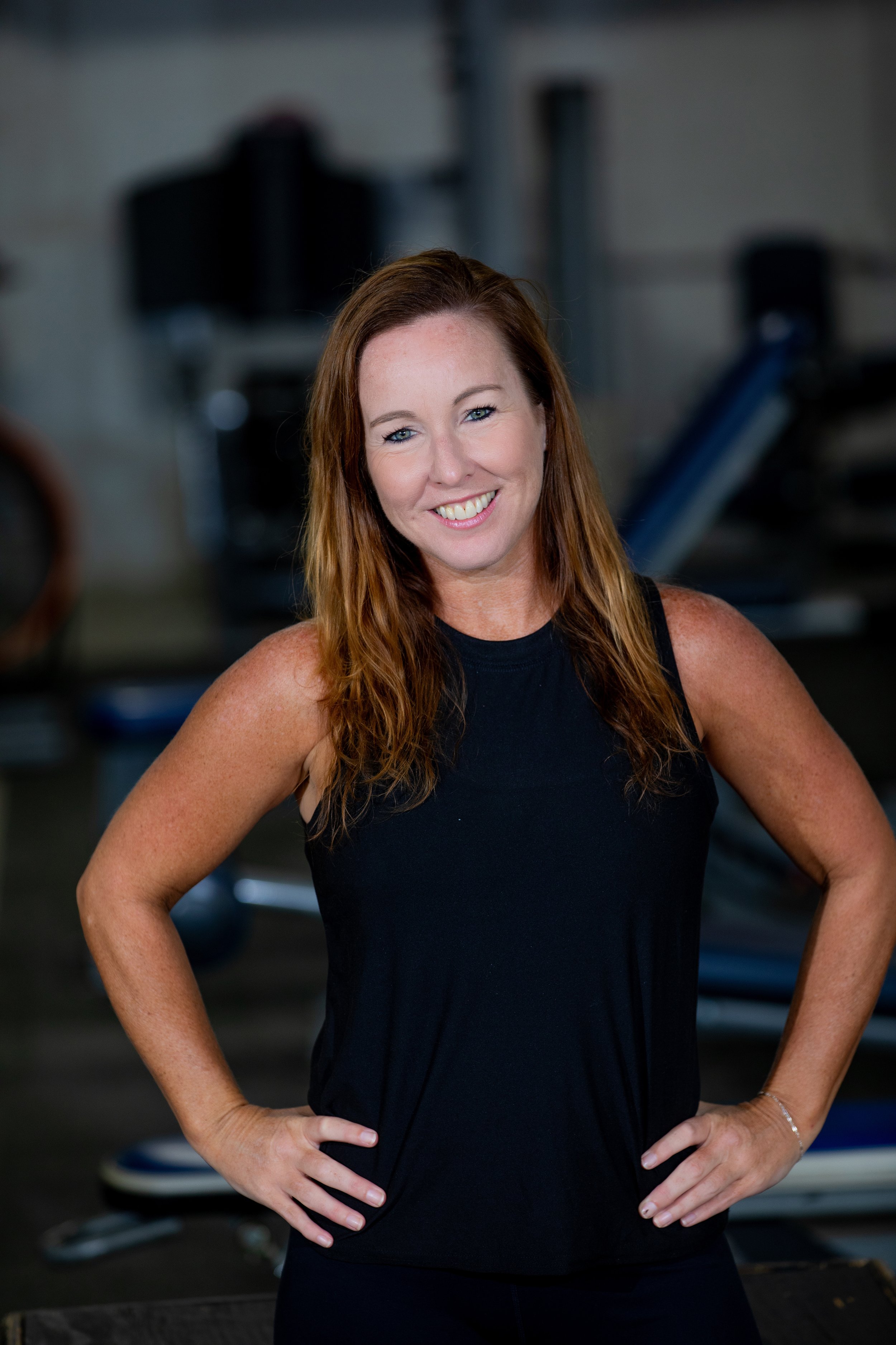 A woman with long red hair smiling, wearing a black sleeveless top, standing with hands on her hips in a gym.