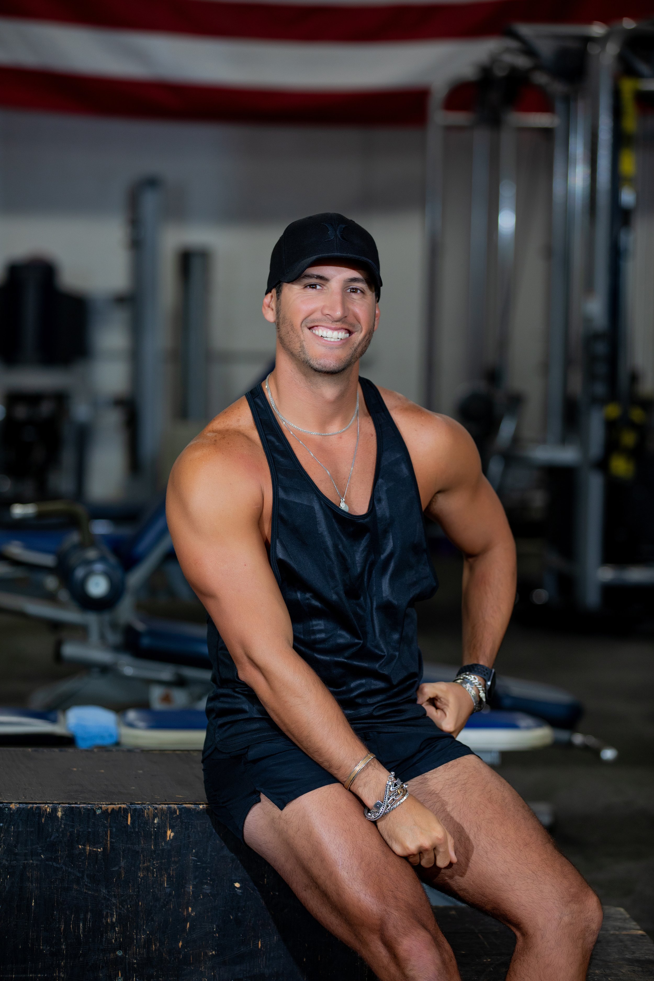 A smiling man in workout attire sitting on a wooden box in a gym, with exercise equipment in the background.