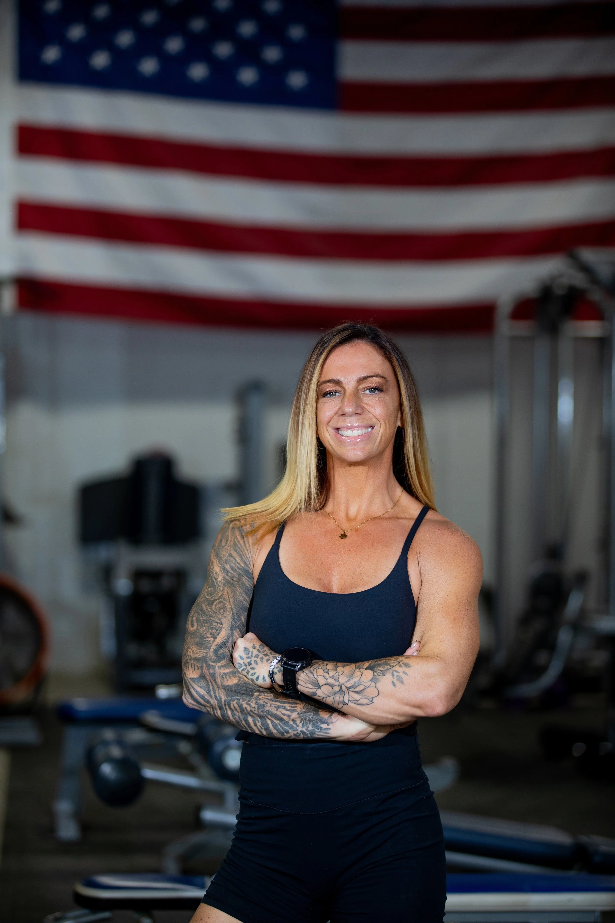 A smiling woman with tattoos on her arms, wearing a black sports top and black shorts, standing with her arms crossed in a gym with an American flag hanging in the background.