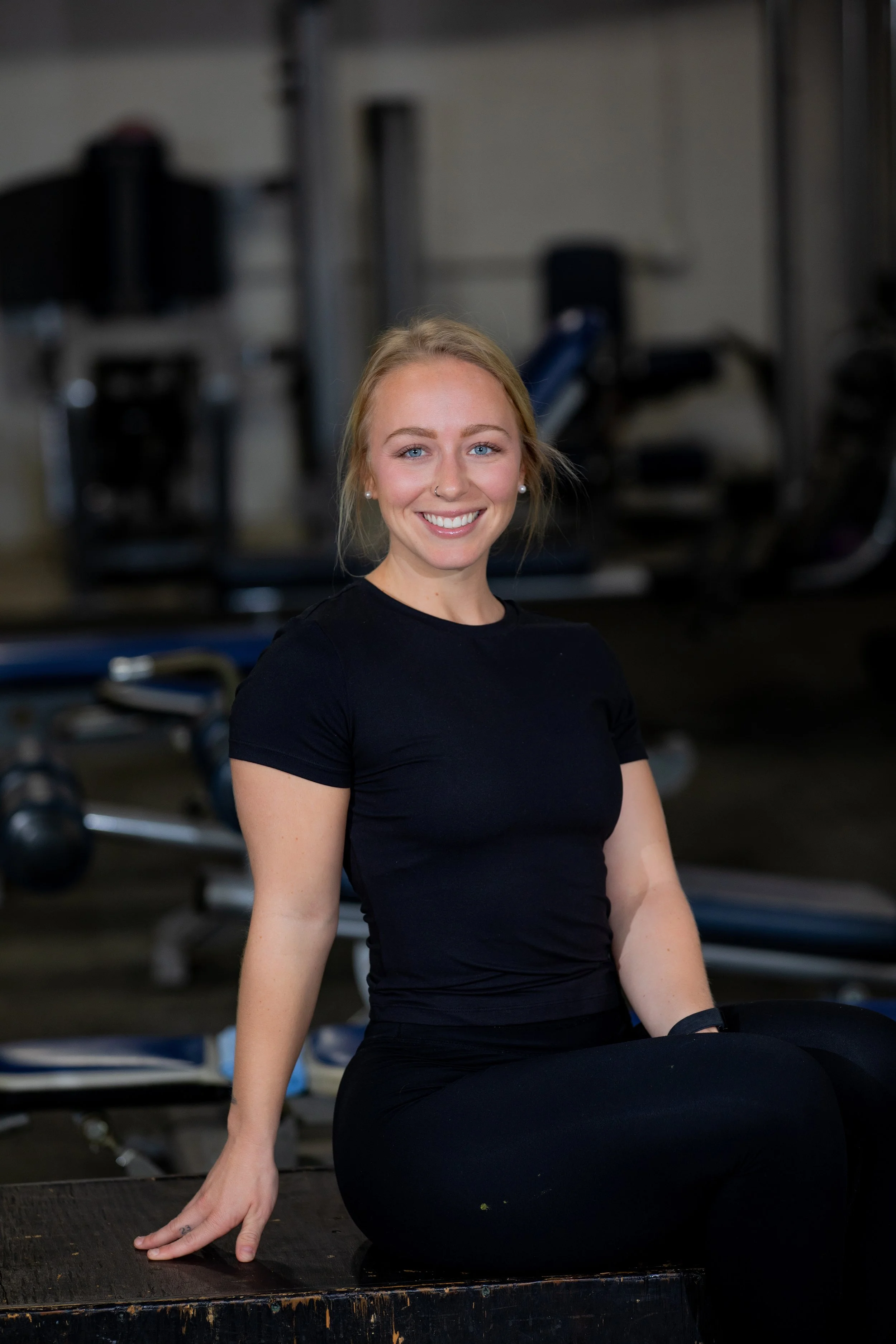 A young woman with blonde hair, wearing a black t-shirt and black leggings, smiling while sitting on a bench in a gym or fitness center.