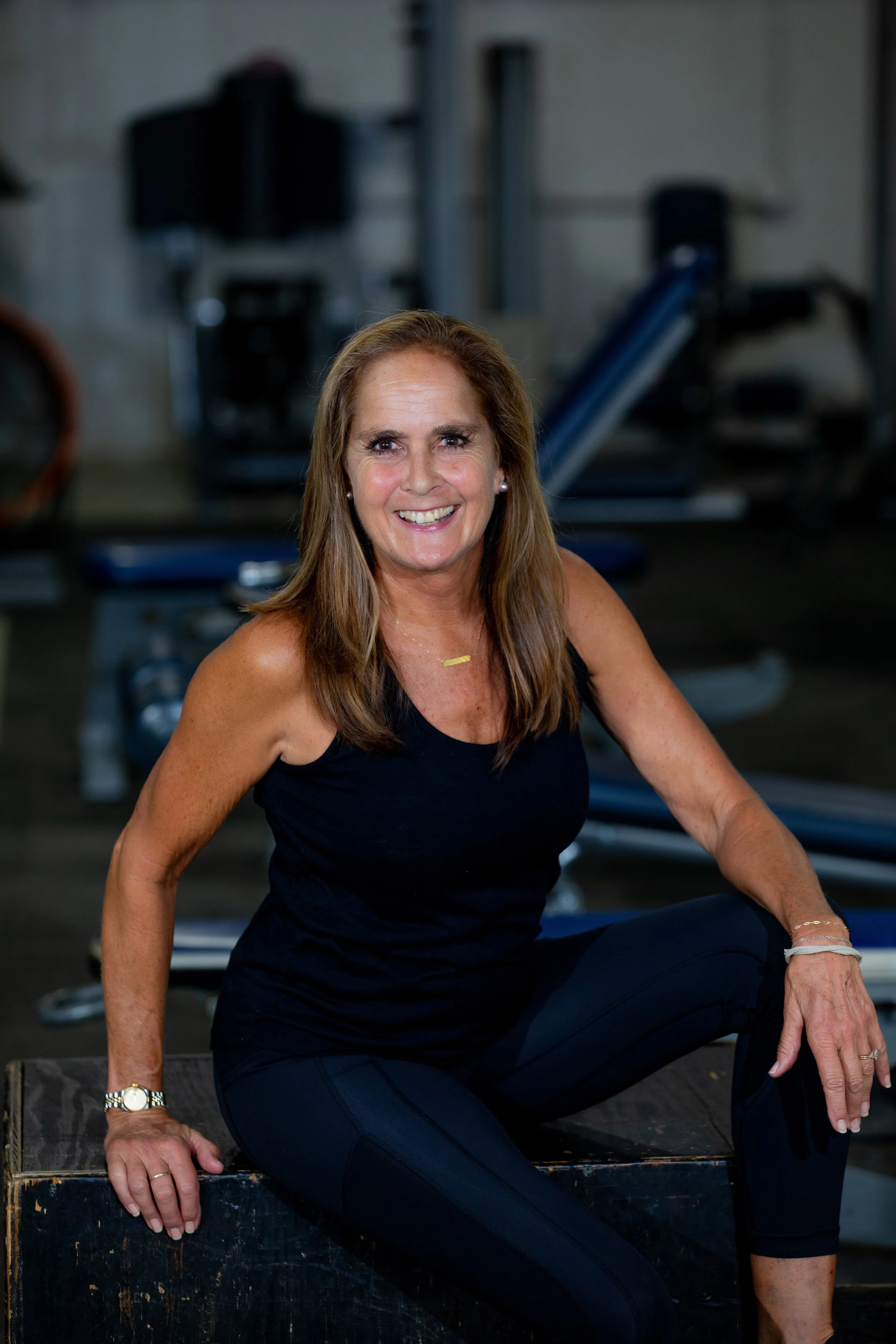 A smiling woman in black workout clothing sitting on a wooden box in a gym with exercise equipment in the background.