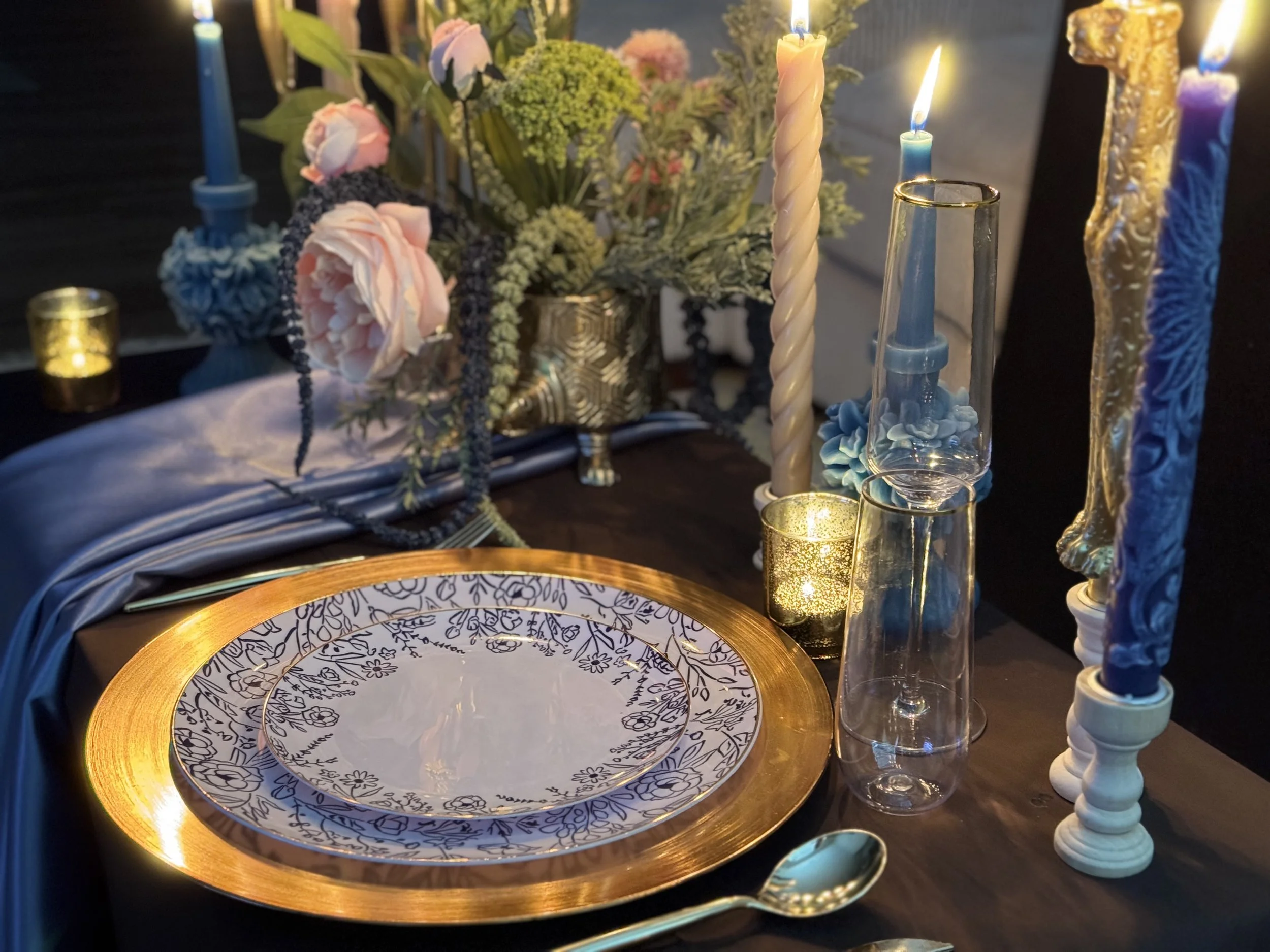 Candlelit dinner table with a blue and white plate on a gold charger, blue taper candles and pink roses