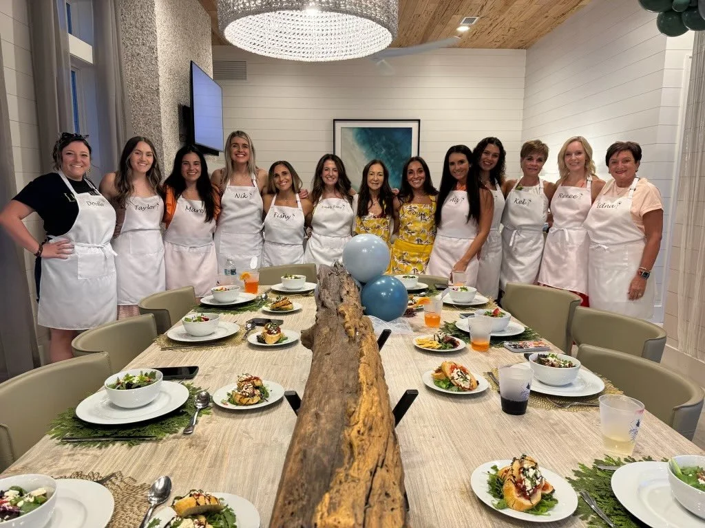 A group of women, wearing aprons with name tags, standing behind a long dining table set with salads, drinks, and appetizers. The women are smiling, and the room is decorated with balloons and artwork on the wall.