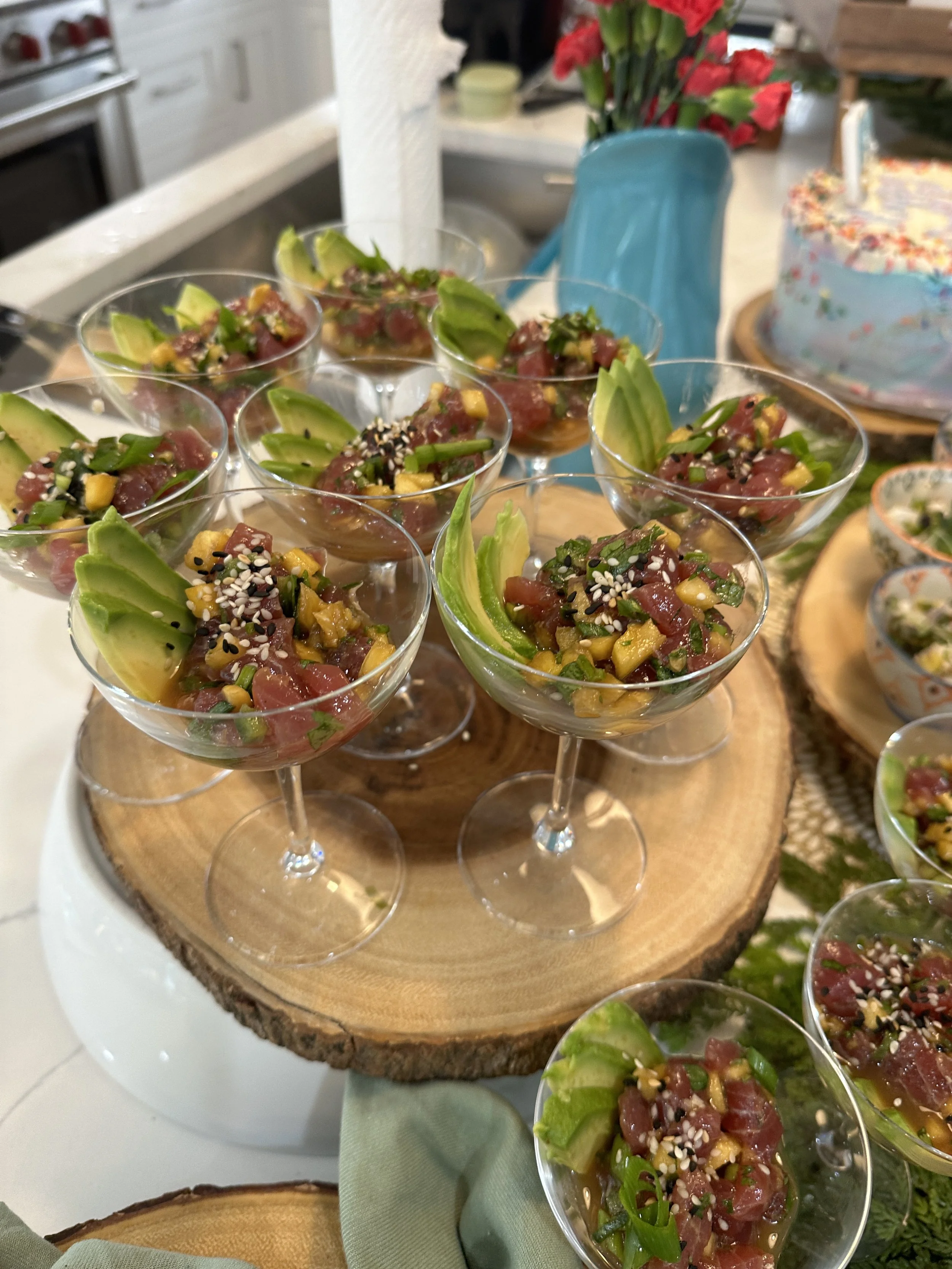 Cocktail glasses filled with tuna tartare, topped with chopped green onions, sesame seeds, and slices of avocado, arranged on a wooden serving stand.
