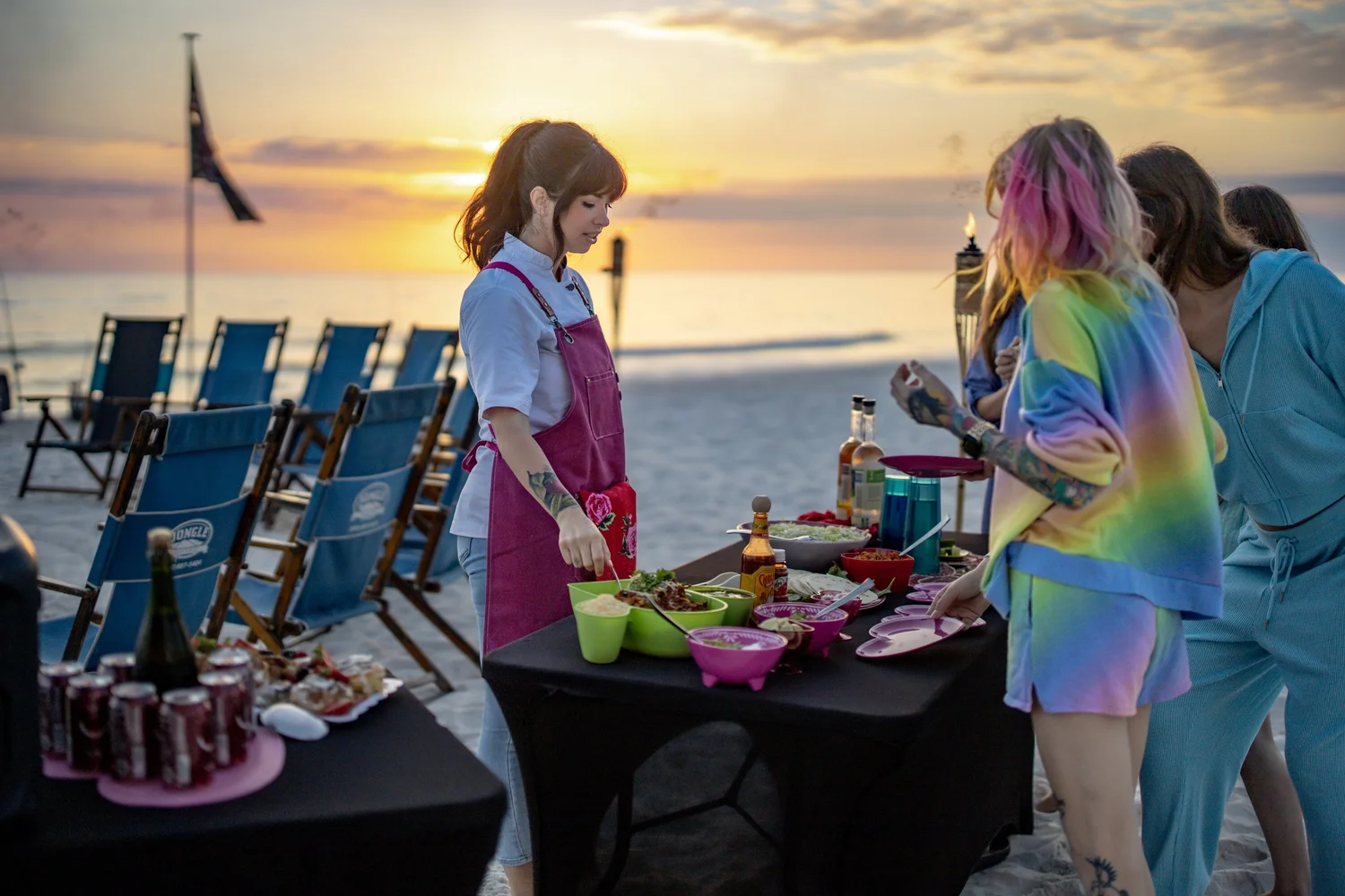 Chef Jojo in her pink apron serving a sunset beach dinner on 30A, with tiki torches and beach chairs