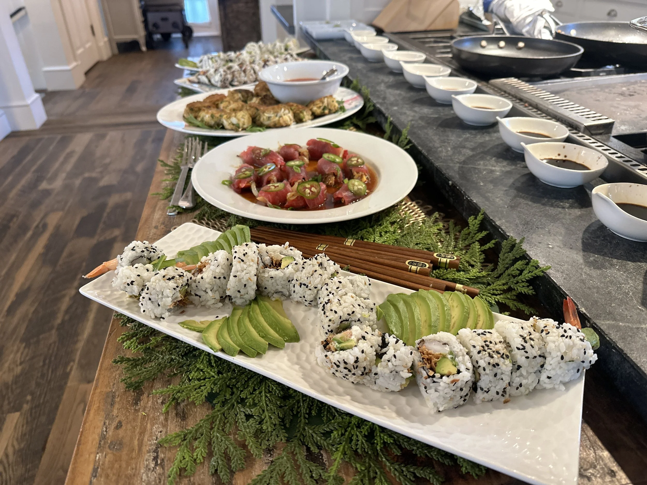 A sushi platter with avocado slices and sushi rolls, arranged on a white rectangular dish, on a wooden table with green foliage. In the background, there are plates of chopped tomatoes with green pepper slices, various dipping sauces, and a kitchen countertop.