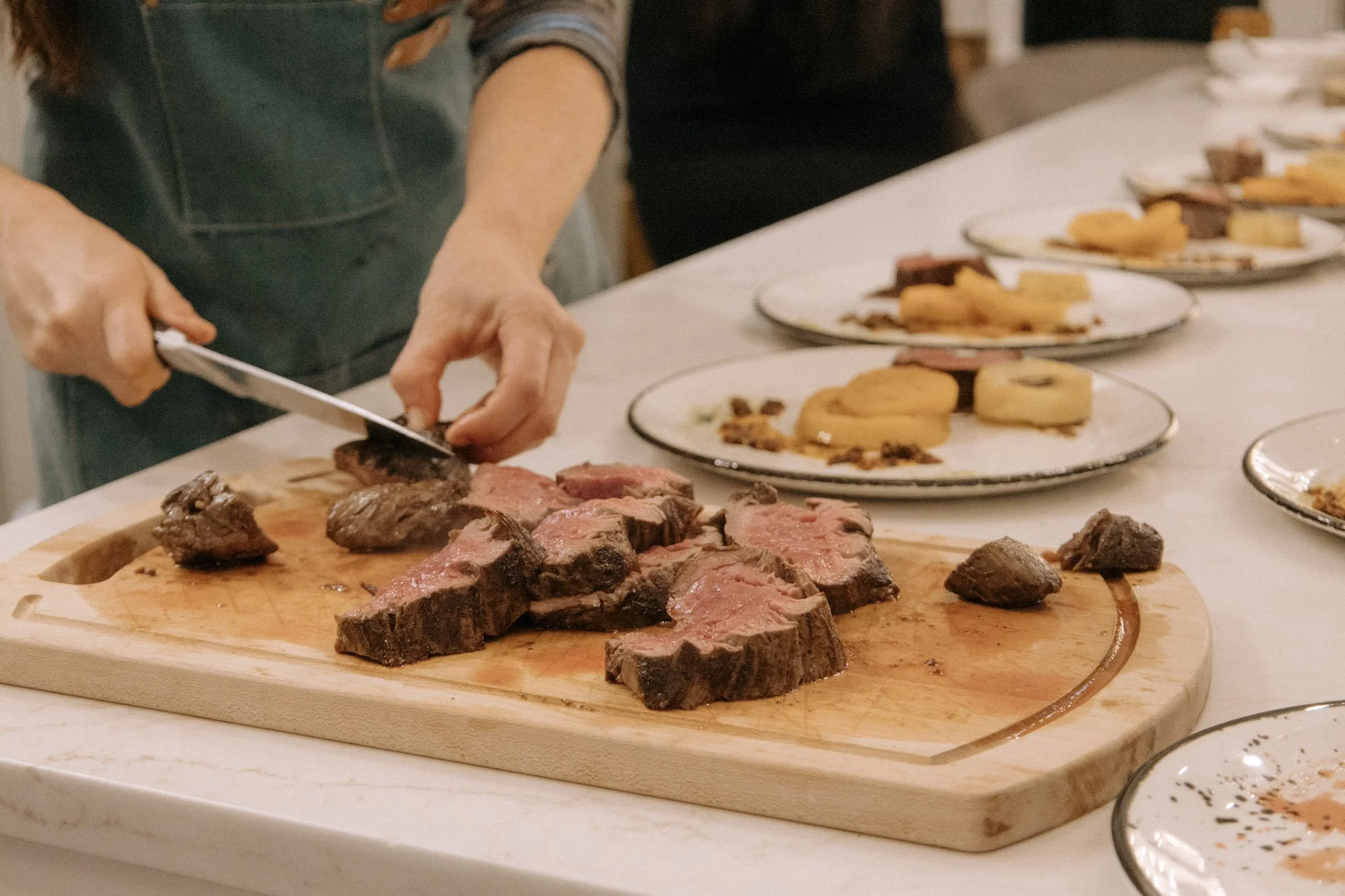 Person slicing cooked steak on a wooden cutting board with plates of assorted foods in the background.