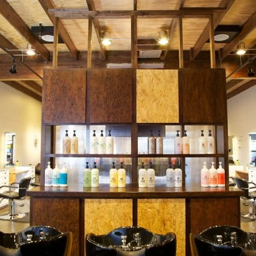 Display of various bottles of hair care products on shelves in a salon, with wooden panels and chairs in the background.