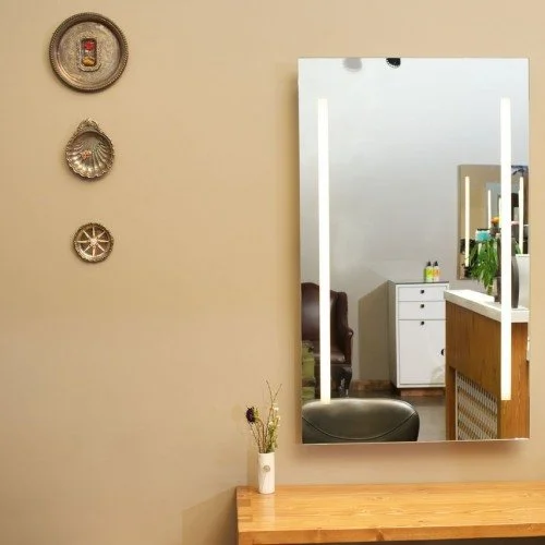 A mirror on a beige wall with three decorative wall hangings above a wooden countertop, reflecting a salon station opposite with a white dresser, a chair, and a plant.