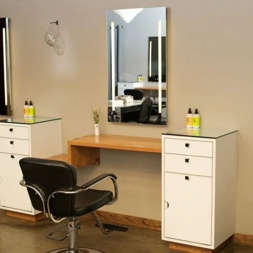 A salon station with a large wall mirror, a wooden countertop, and two white storage cabinets with bottles on top. There is a black salon chair in front of the mirror and a small vase with flowers on the counter.