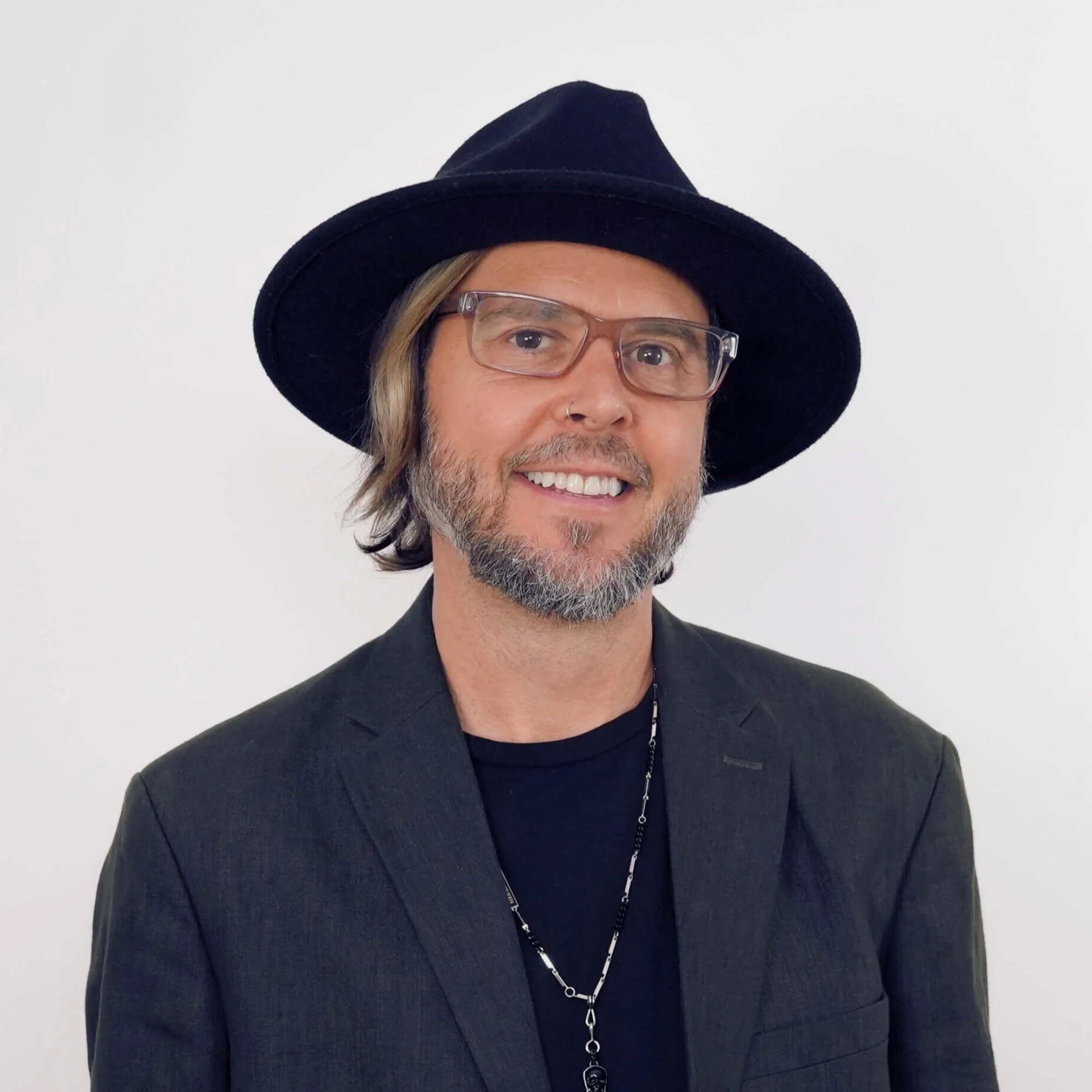 A man with glasses, a beard, and a mustache wearing a black fedora hat, a black blazer, and a black shirt, smiling at the camera against a plain white background.