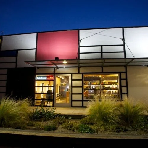 A modern storefront with large glass windows, illuminated from inside, featuring shelves with products, and surrounded by landscaped plants at night.