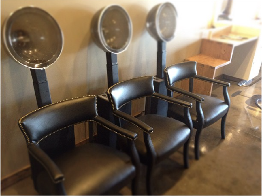 Three black salon chairs with hair dryers attached behind them in a row, positioned against a beige wall in a salon.