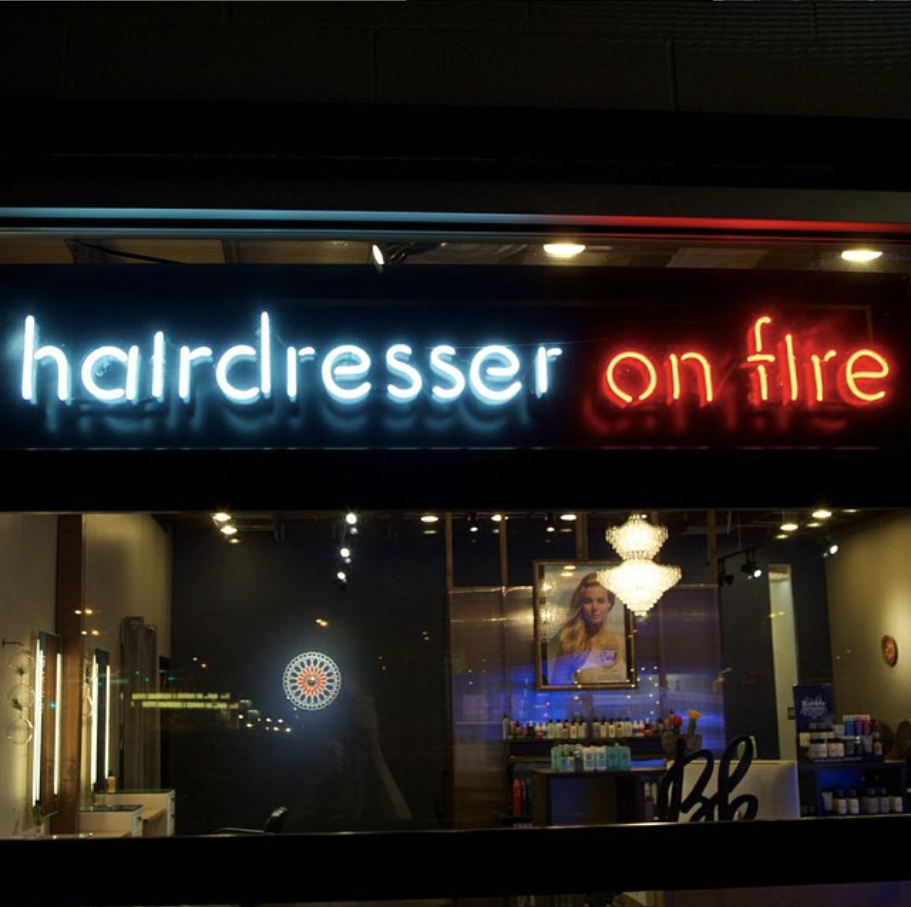 Neon sign reading 'hairdresser on fire' with a salon interior visible through the window, including a chandelier, framed photo, and various hair products.