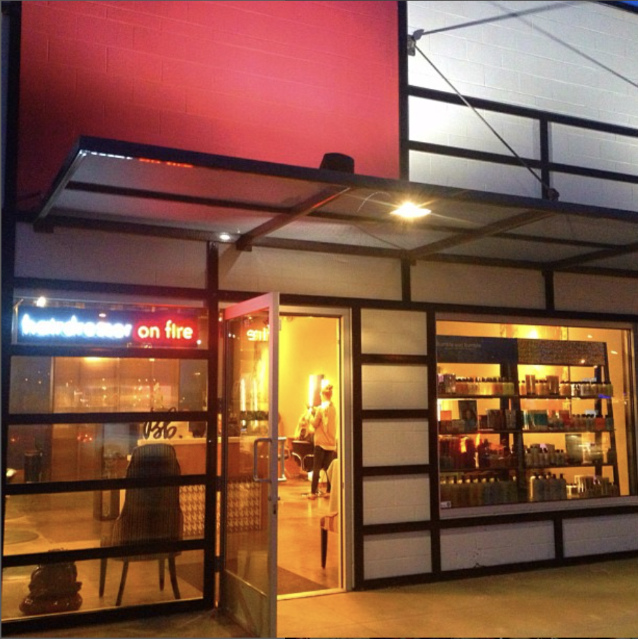Exterior of a store with a glass door and shelves filled with products, lit with warm yellow lighting, and a neon sign that says 'hairdresser on fire' in red, with the storefront showing a red and white modernist design.
