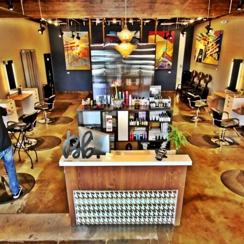 Interior of a modern hair salon with styling stations, mirrors, chairs, and colorful abstract artwork on the walls. A reception desk with beauty products and decorative items is in the foreground.