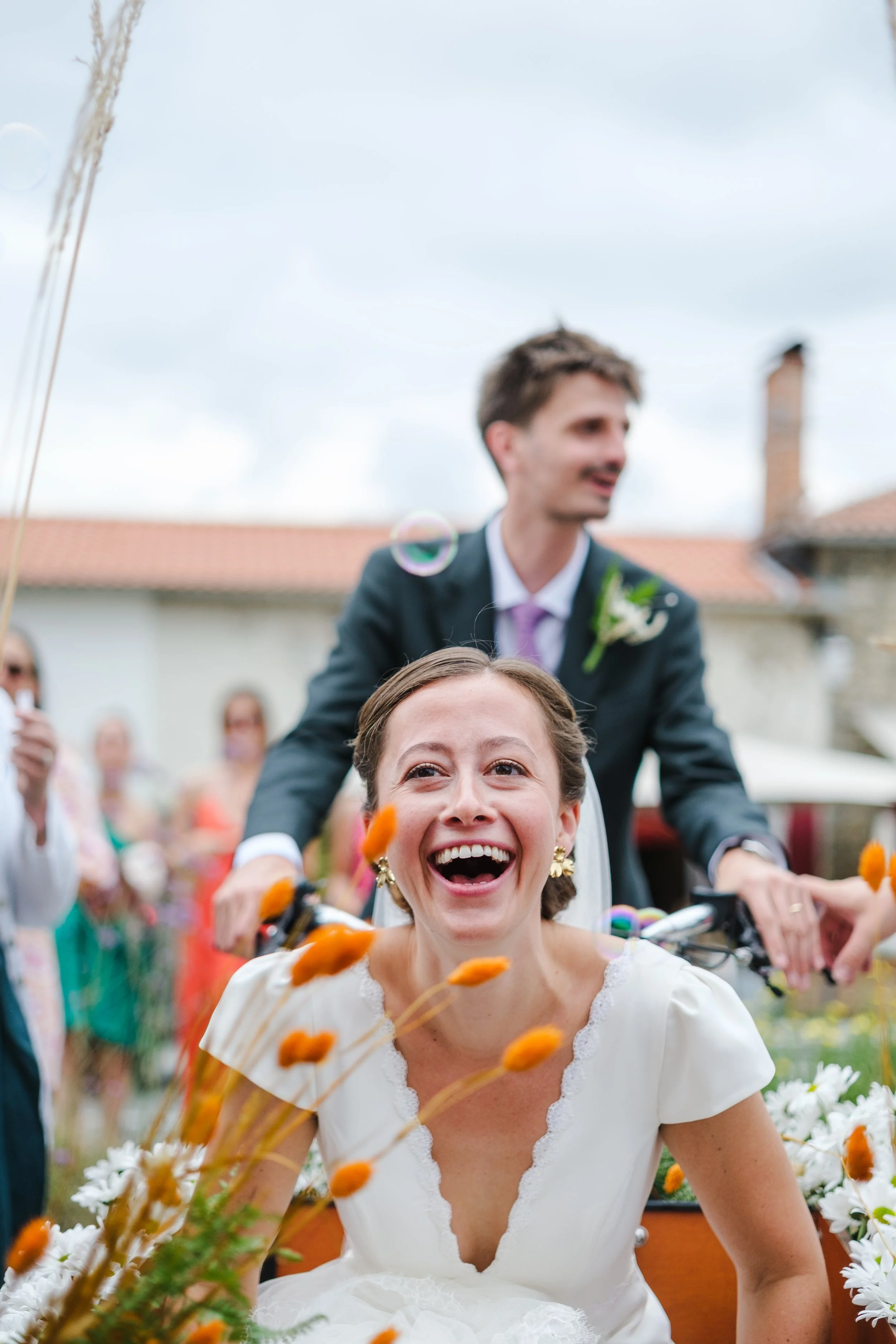 Une femme heureuse en robe blanche durant une célébration ou un mariage, avec un groupe de personnes en arrière-plan, décorée de fleurs et dans un environnement extérieur.