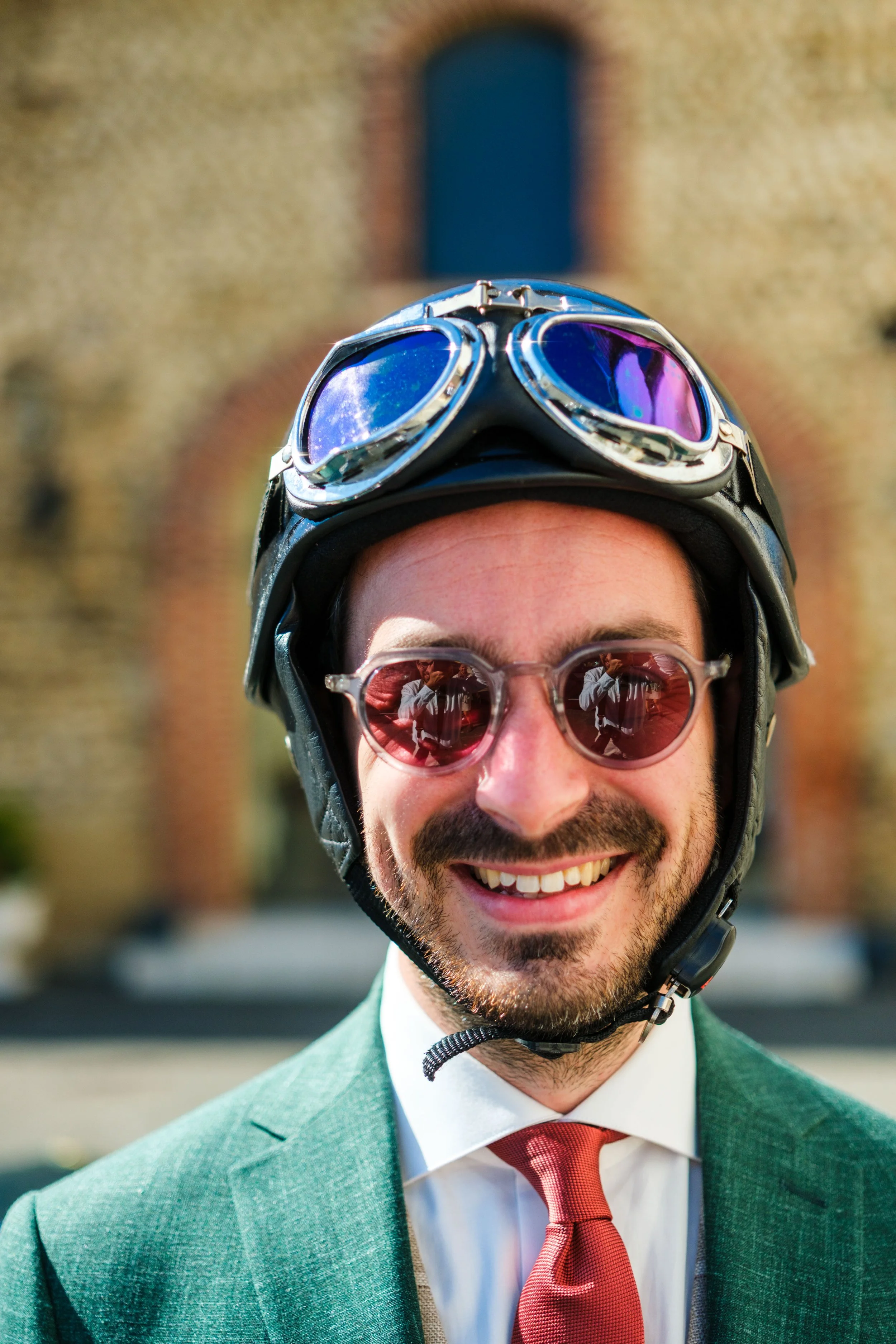 Un homme souriant porte un casque de moto avec des lunettes de soleil réfléchissantes. Il porte un costume vert avec une chemise blanche et une cravate rouge. L'arrière-plan montre un bâtiment ancien en briques.