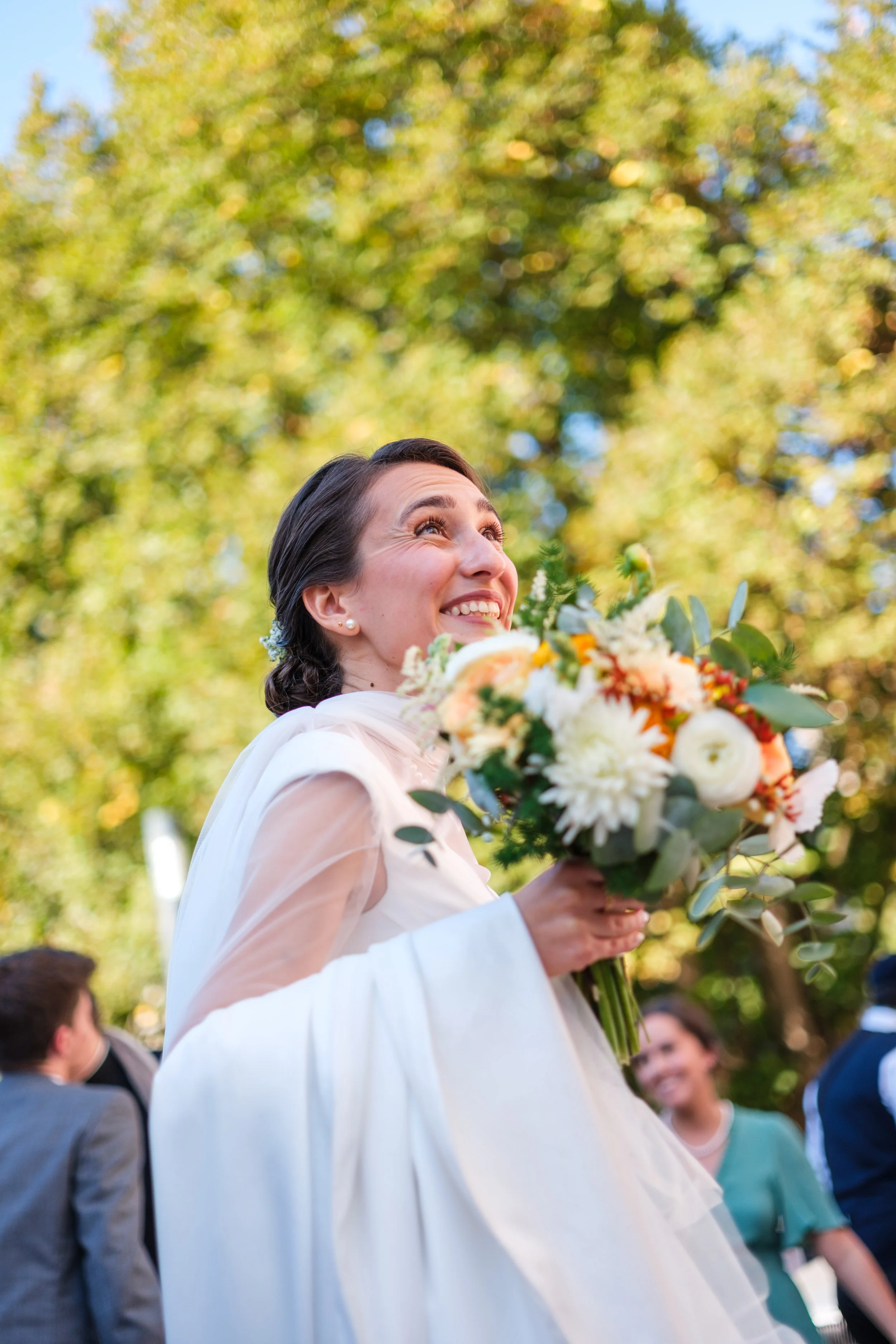 Une jeune femme souriante en robe blanche, tenant un bouquet de fleurs, lors d'une célébration en plein air, avec des personnes en arrière-plan.